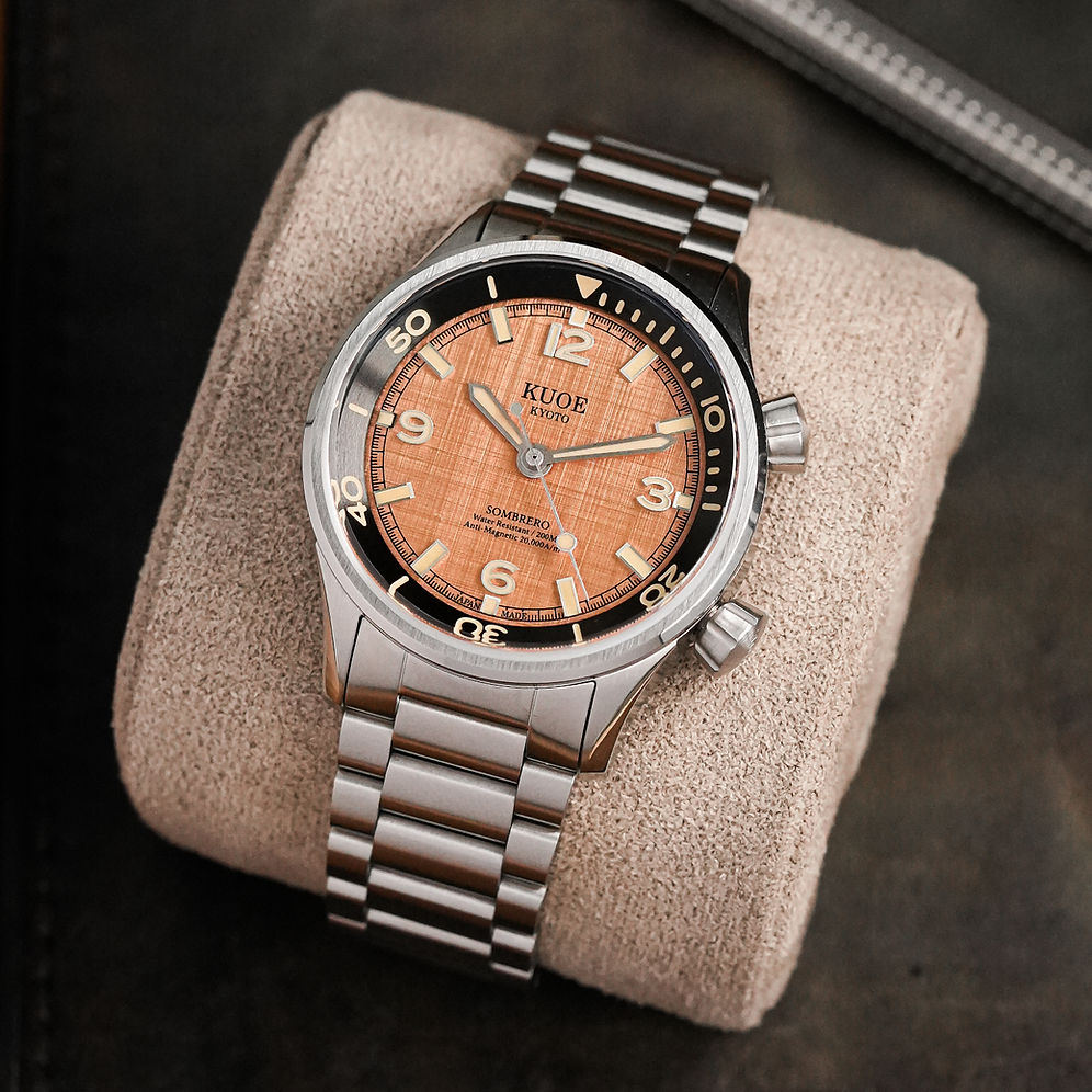 A Kuof wristwatch with a stainless steel link bracelet and a copper-orange dial, resting on a beige textured cushion over a dark background.