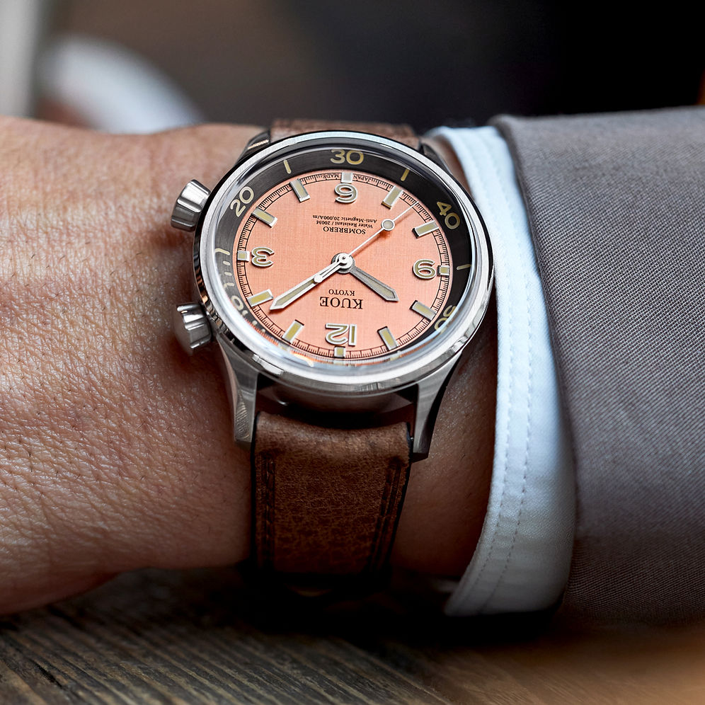 A close-up of a wrist wearing a Rolex Submariner Date watch with a distinctive peach-orange dial, black bezel, and a brown leather strap, styled with a white dress shirt cuff and gray suit jacket.