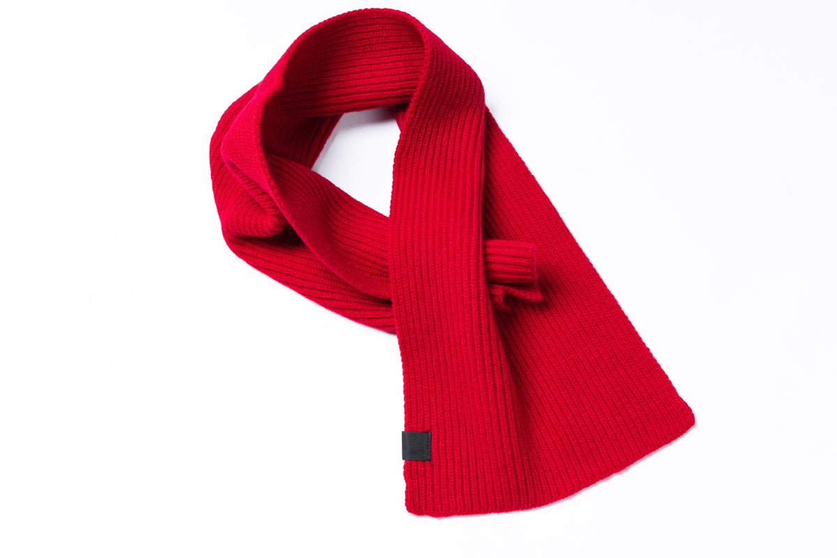 A bright red ribbed knit scarf with a small black square tag near the bottom, styled in a loop on a plain white background.