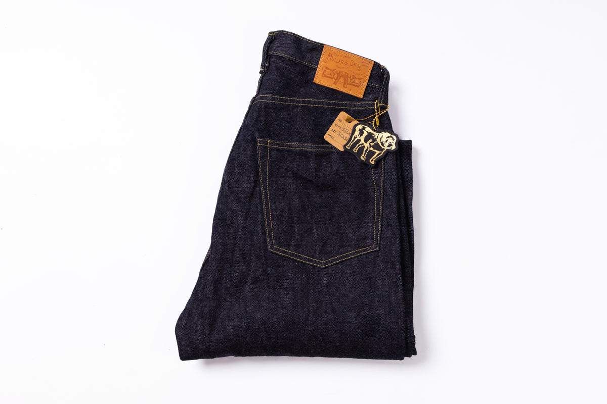 A folded pair of dark indigo denim jeans with a visible orange leather waistband patch and a paper brand tag, photographed on a clean white background.