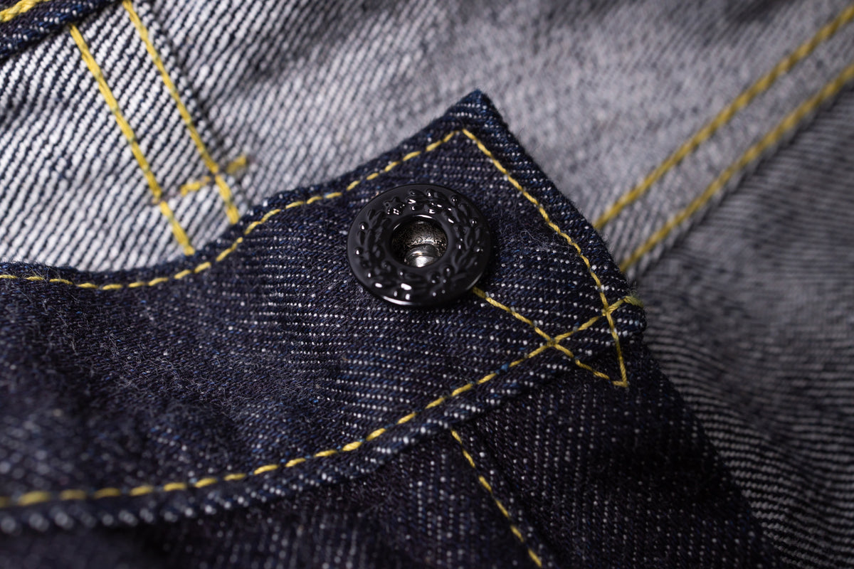 Close-up of dark indigo denim fabric with yellow contrast stitching and a black metal snap button.