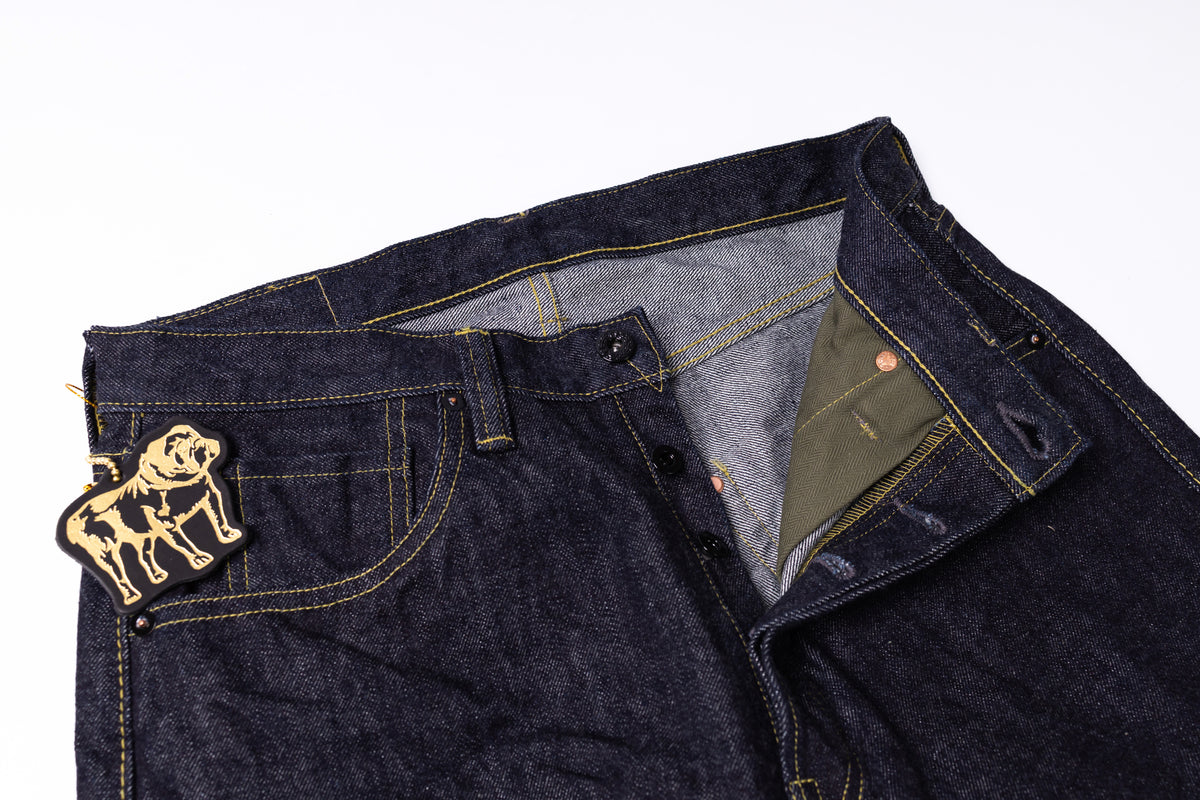 Close-up of dark indigo denim jeans with contrast yellow stitching, a gold embroidered bulldog patch on the pocket, and an olive green inner pocket lining, shown on a white background.