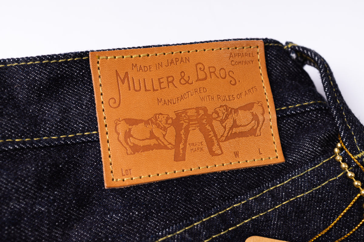Close-up of a dark indigo denim waistband with a tan leather patch sewn onto the inside back waist. The patch is embossed with the Muller & Bros.