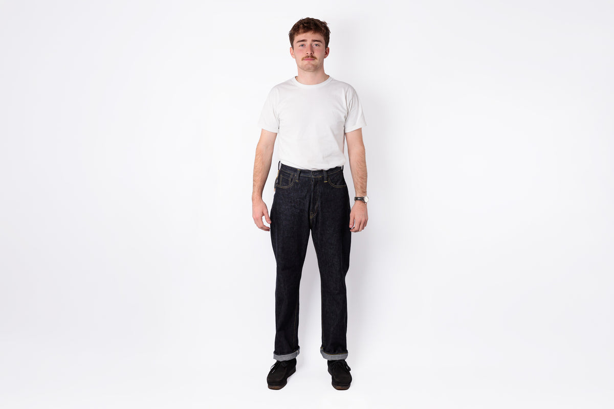 A man standing against a plain white studio background, wearing a plain white short-sleeve t-shirt and dark blue jeans.