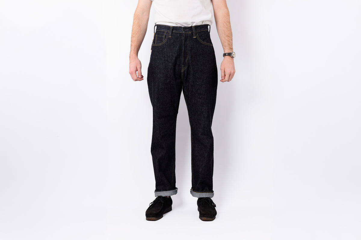 A person wearing dark indigo denim jeans with rolled cuffs, paired with a white t-shirt and black shoes, standing against a plain white studio background.