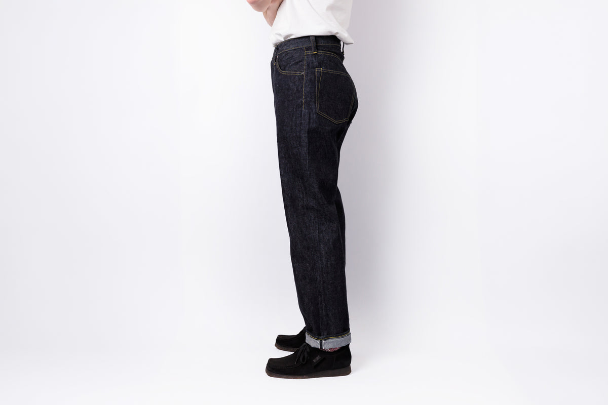 A side profile view of a person wearing dark indigo denim jeans, a white shirt, and black shoes against a plain white studio background.