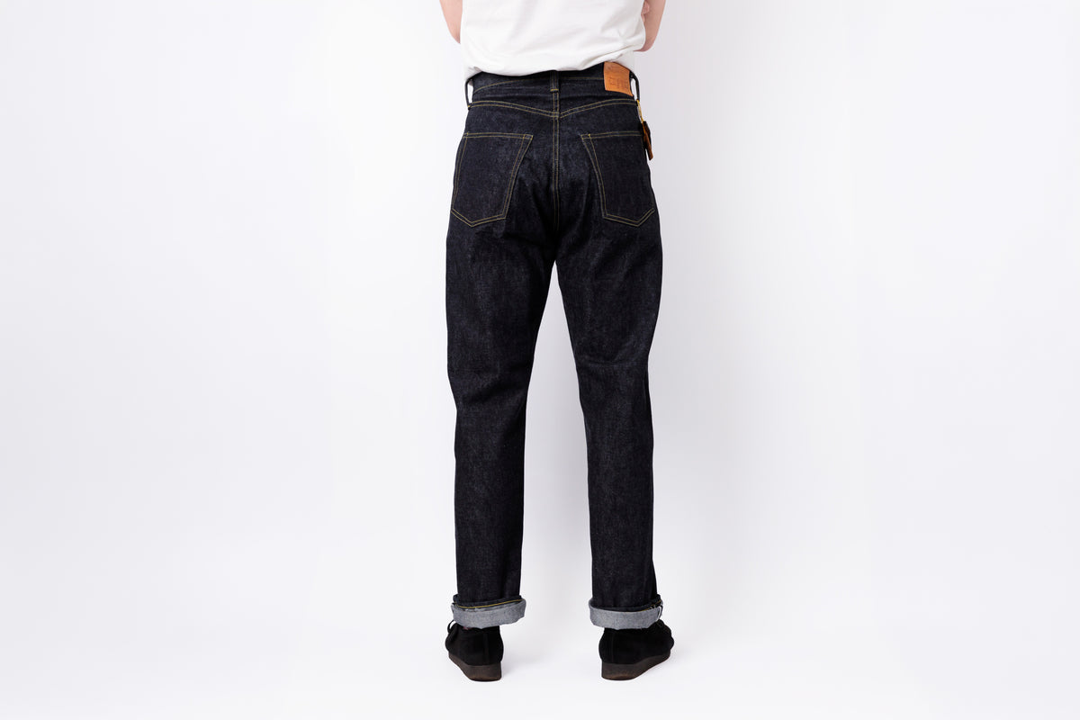 Rear view of a person wearing dark indigo denim jeans with a white shirt and black shoes, standing against a plain white studio background.