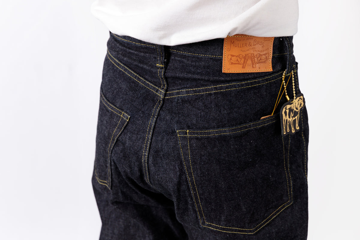 Close-up back view of a person wearing dark indigo denim jeans with a white shirt tucked in. A tan leather patch is visible at the back waistband, and a small gold-tone animal charm hangs from the back pocket.