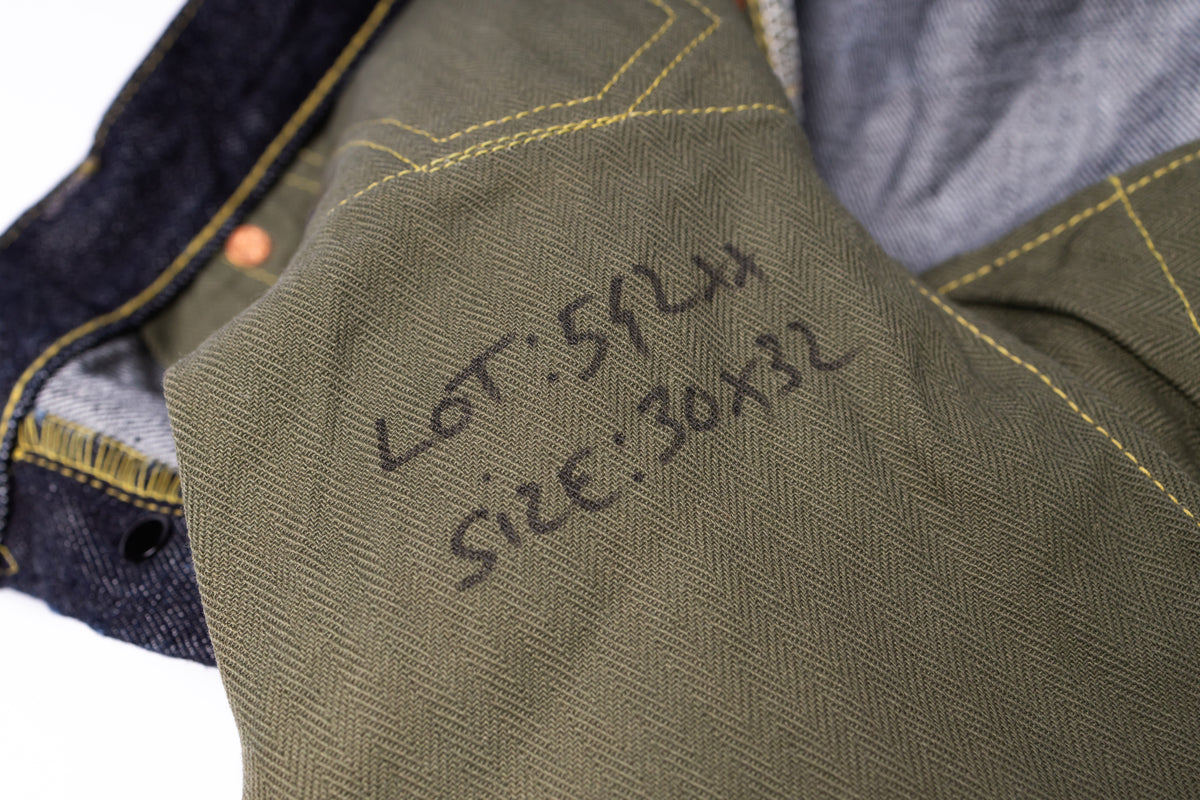 Close-up of olive green herringbone fabric with yellow stitching and handwritten black ink text reading 'LOT: 59LM' and 'SIZE: 30x32', next to a dark denim material.