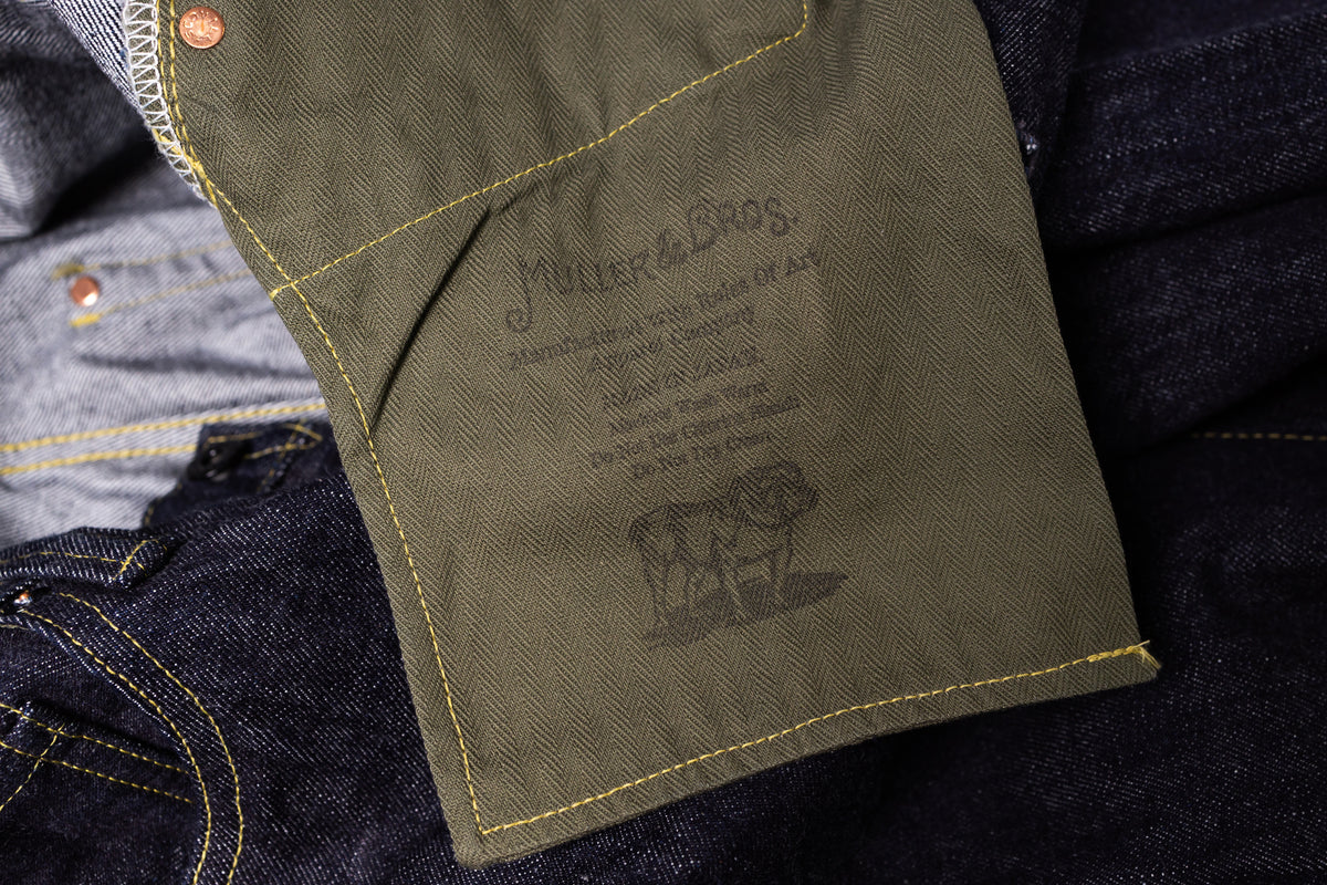 Close-up of an olive green denim patch pocket on a pair of dark indigo jeans, showing yellow contrast stitching and an interior label printed with the brand 'Mutter & Bros.' and a cow logo.