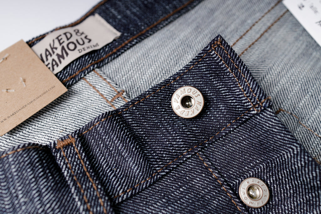 Close-up of denim fabric with button and 'Naked & Famous' label.