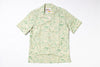 A short-sleeve button-up camp shirt with a light green and beige tropical landscape print, laid flat against a white background.