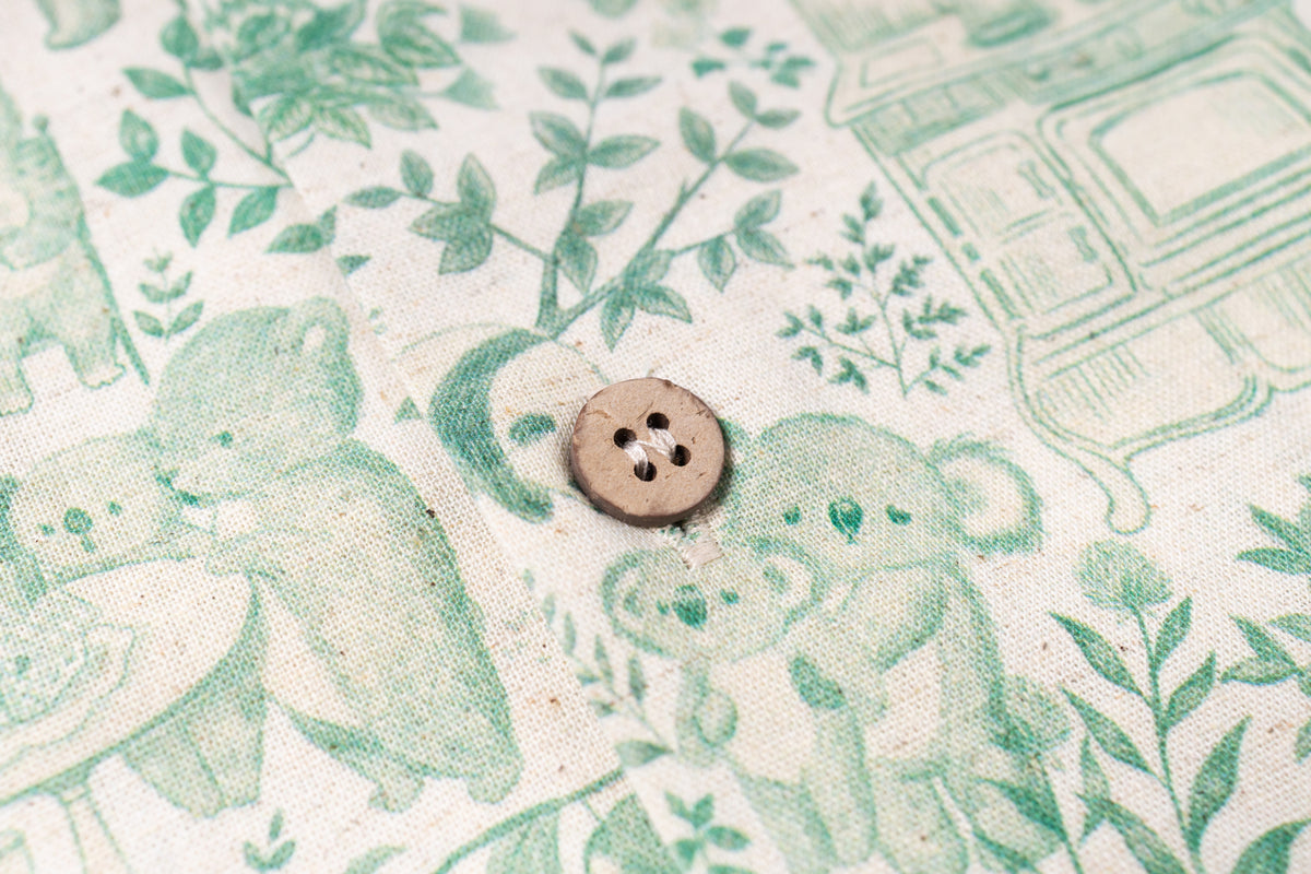 A close-up of a light brown four-hole button resting on a cream-colored fabric with a green botanical and animal print.