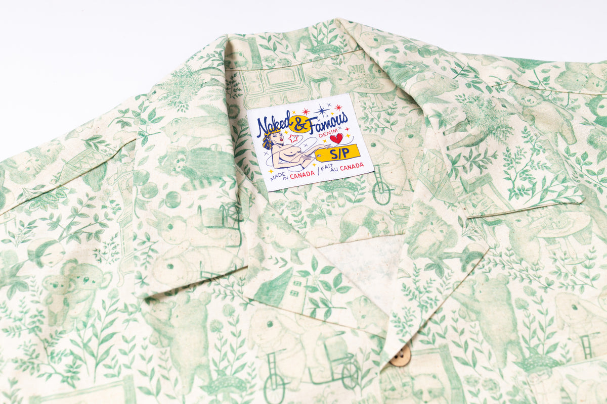 A close-up of a short-sleeve button-up shirt with a light beige and green botanical pattern, featuring a visible 'Natural & Famous' brand label.
