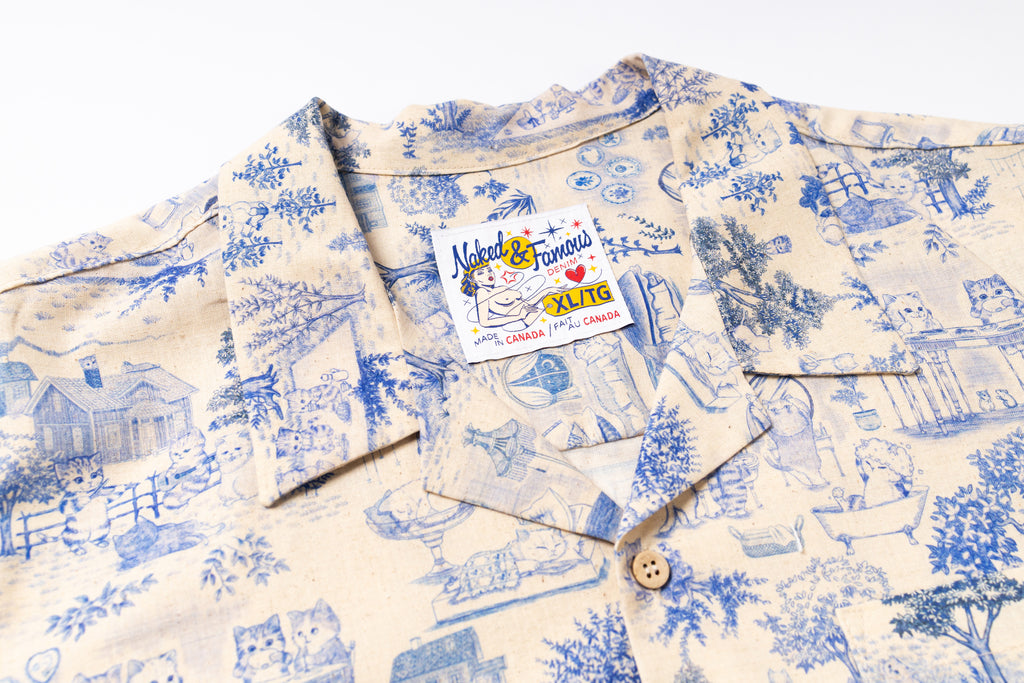A close-up of a short-sleeve button-up shirt with a blue pattern on a cream-colored background. The shirt features a visible brand label for 'Natural & Famous ' and a wooden button at the collar.
