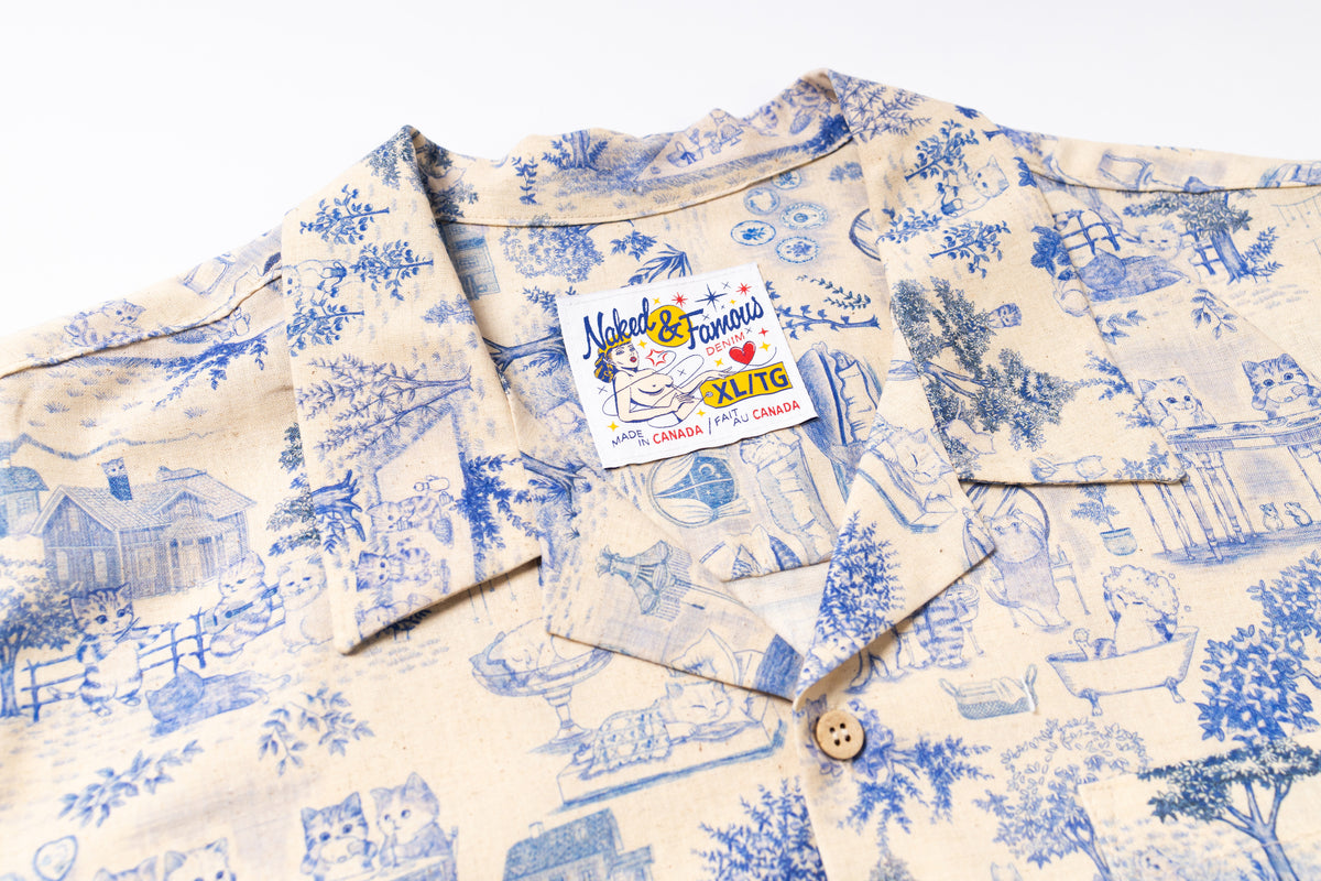 A close-up of a short-sleeve button-up shirt with a blue pattern on a cream-colored background. The shirt features a visible brand label for 'Natural & Famous ' and a wooden button at the collar.