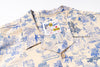 A close-up of a short-sleeve button-up shirt with a blue pattern on a cream-colored background. The shirt features a visible brand label for 'Natural & Famous ' and a wooden button at the collar.