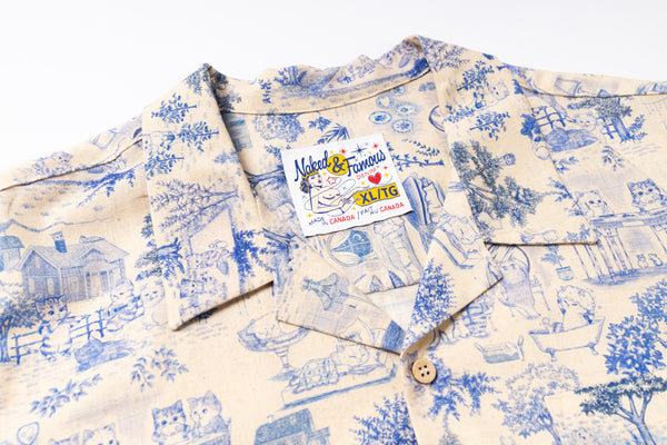 A close-up of a short-sleeve button-up shirt with a blue pattern on a cream-colored background. The shirt features a visible brand label for 'Natural & Famous ' and a wooden button at the collar.