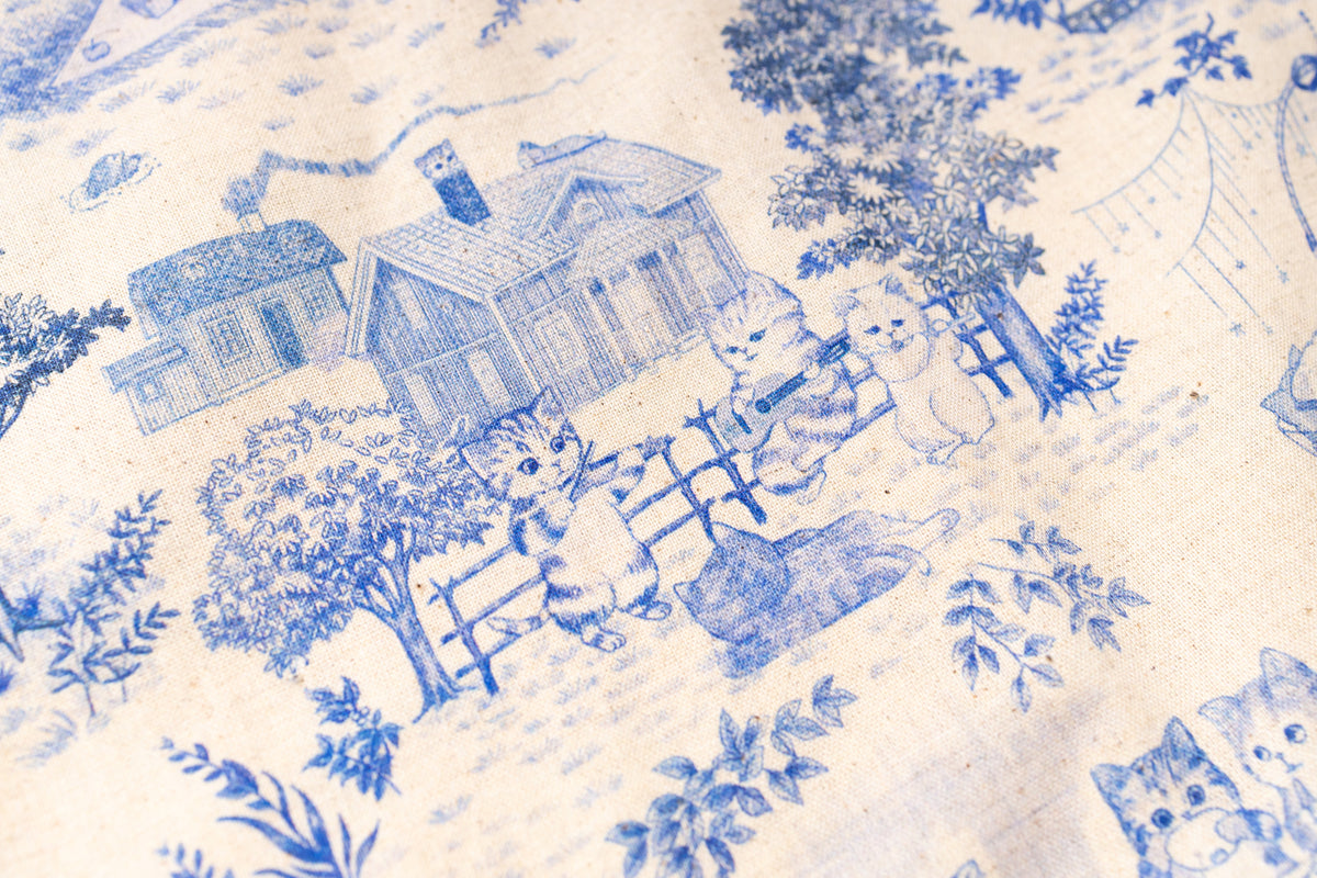 Close-up of a blue and cream toile pattern featuring cats, houses, trees, and foliage on a light fabric surface.