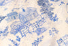 Close-up of a blue and cream toile pattern featuring cats, houses, trees, and foliage on a light fabric surface.