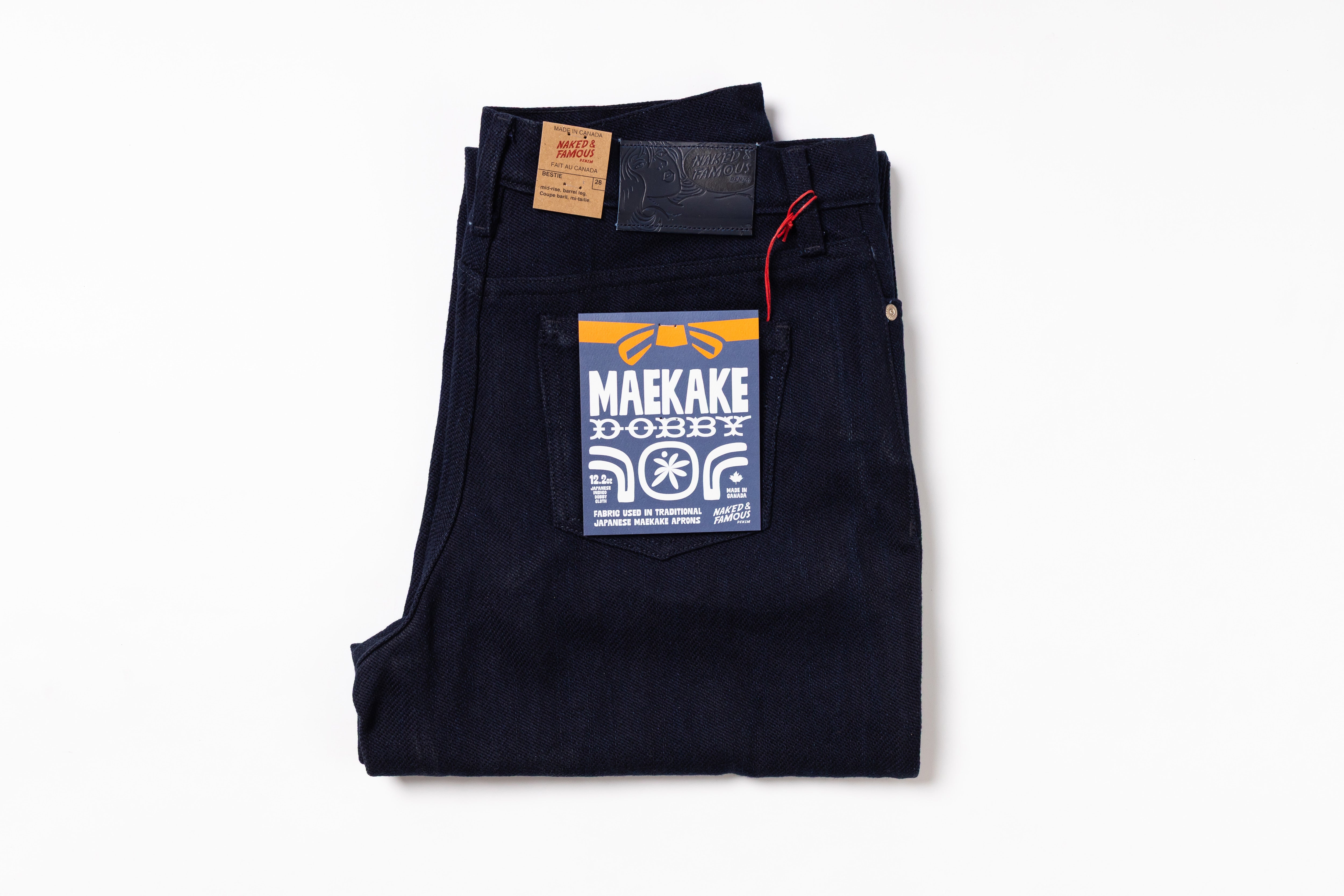 A pair of folded dark indigo denim jeans with a prominent blue, yellow, and white 'MAKAKE DOBBY' patch on the back pocket, shown on a clean white background.