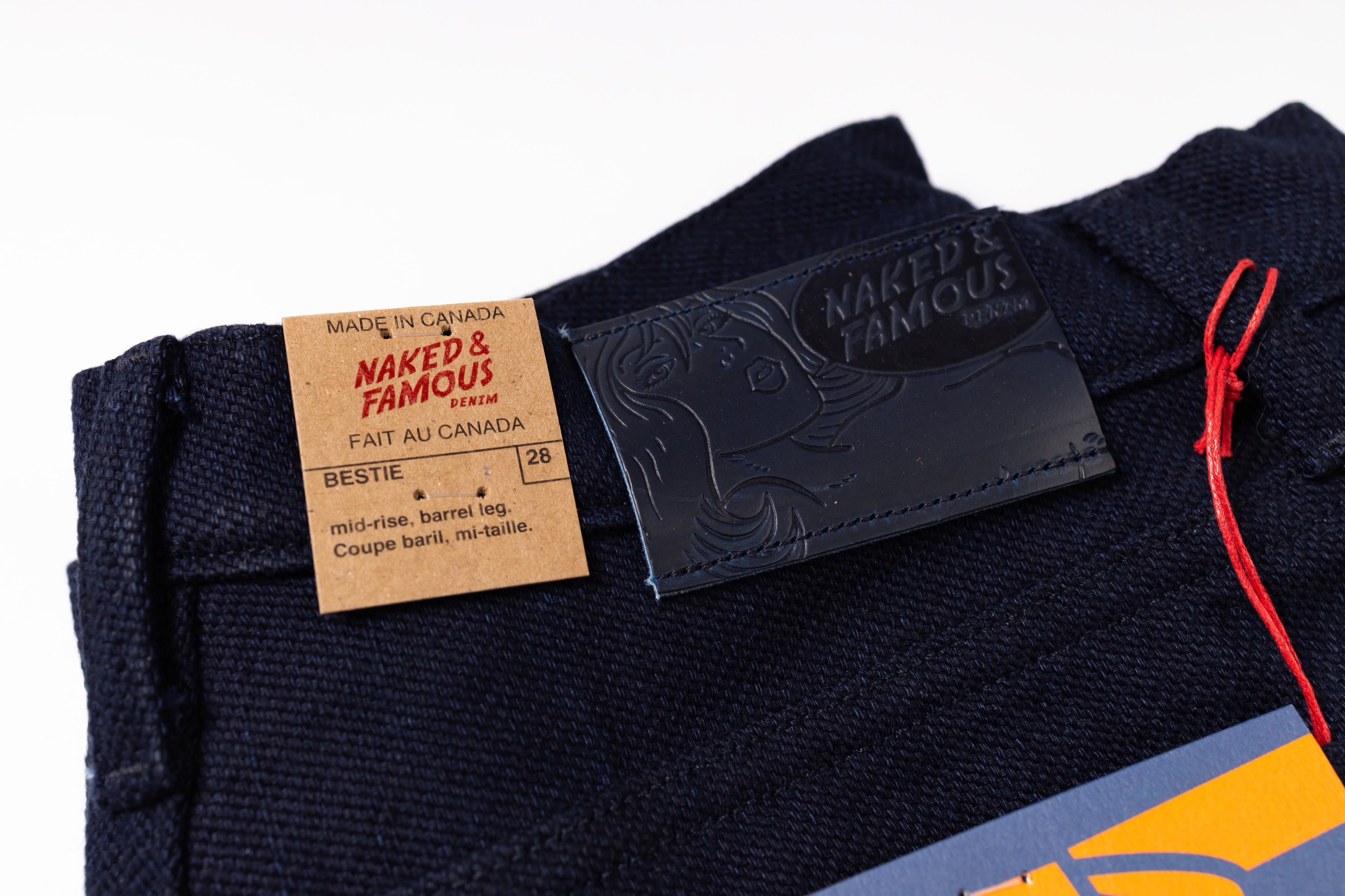 Close-up of the waistband of dark navy blue denim jeans, showing a tan paper brand label reading Naked & Famous and a black leather patch with the same brand name, on a white background.