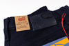 Close-up of the waistband of dark navy blue denim jeans, showing a tan paper brand label reading Naked & Famous and a black leather patch with the same brand name, on a white background.