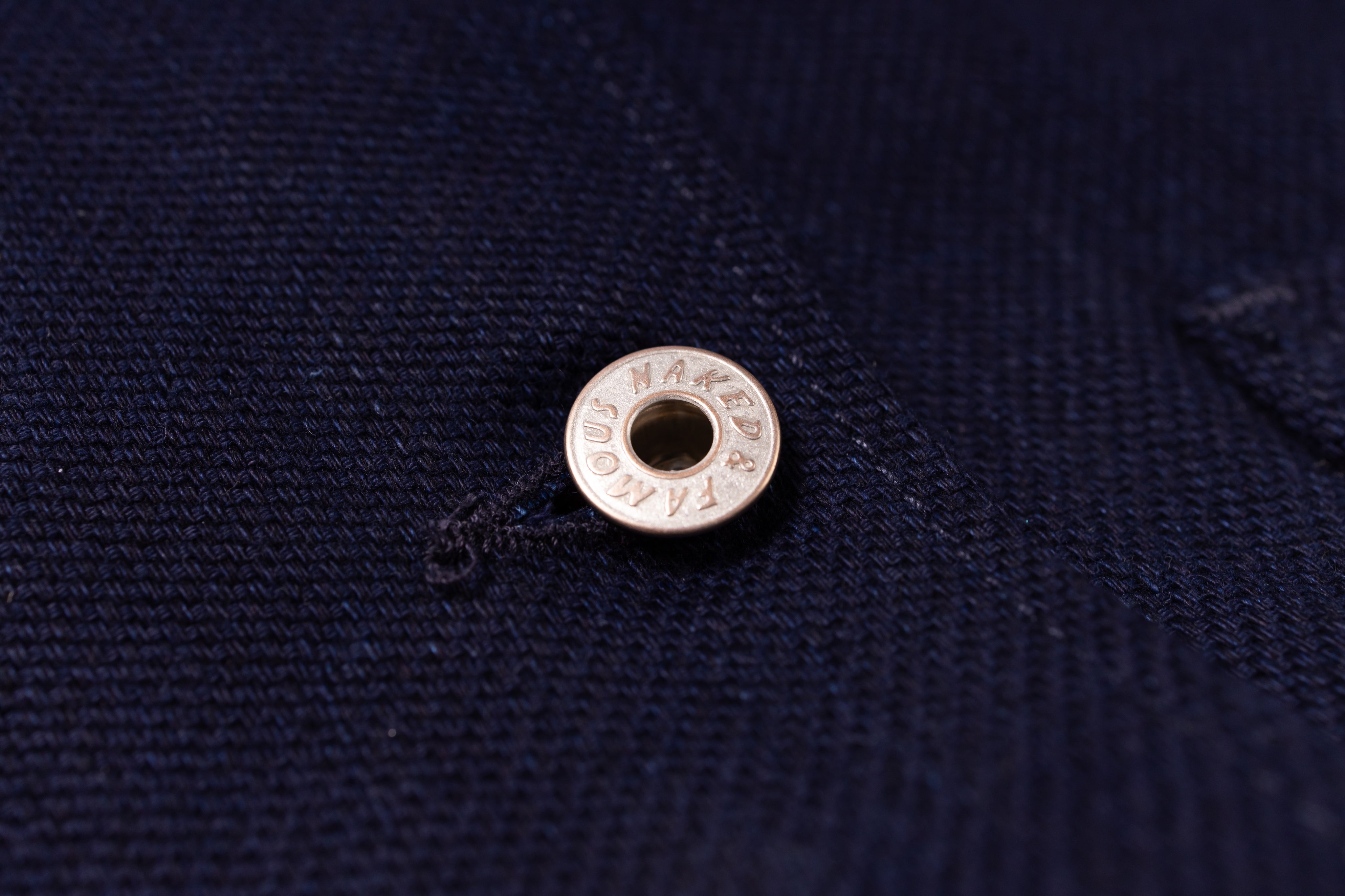 A close-up of a silver metal snap button with the brand name printed on int, set against a dark navy blue textured fabric background.