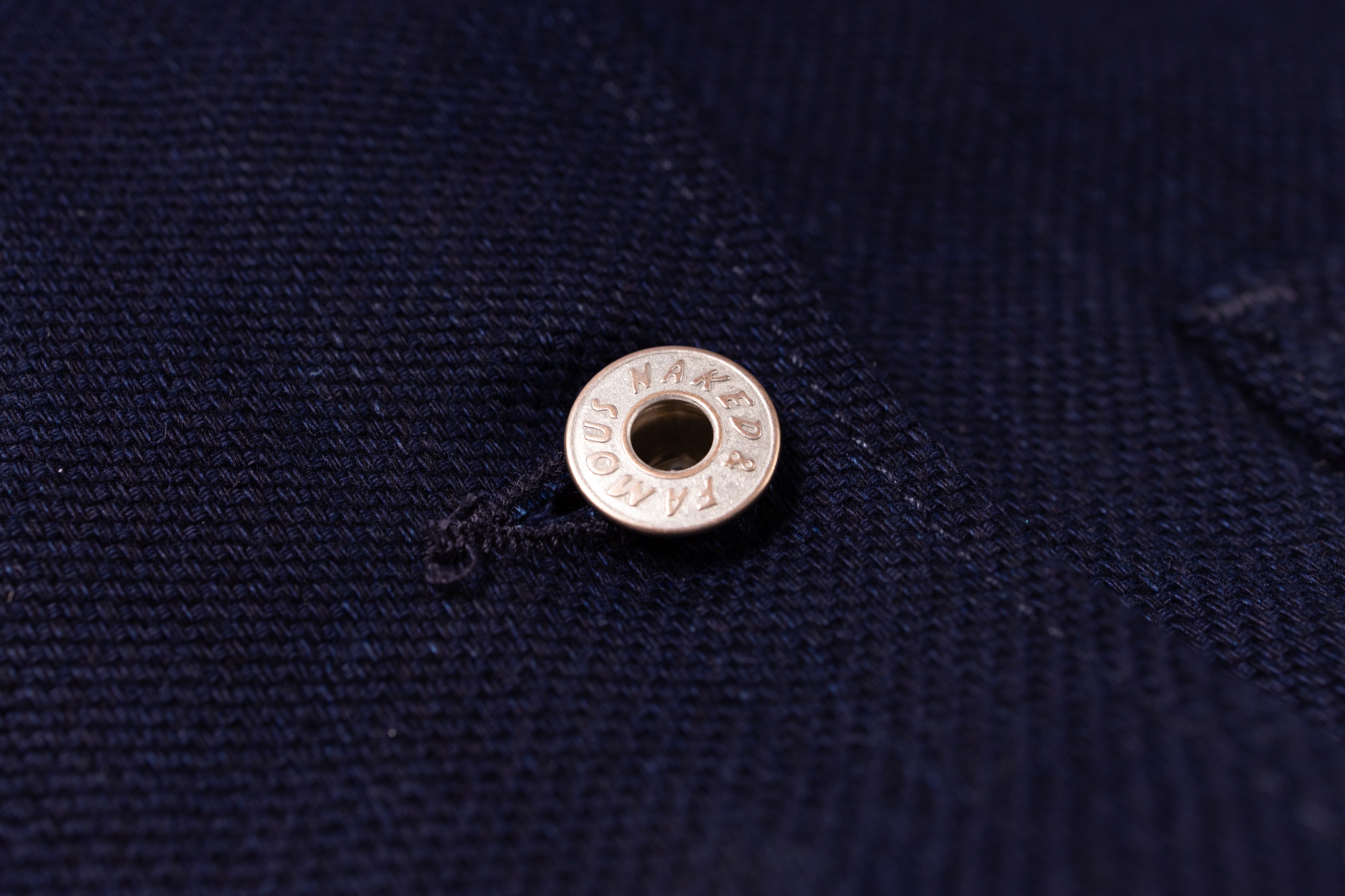A close-up of a silver metal snap button with the brand name printed on int, set against a dark navy blue textured fabric background.