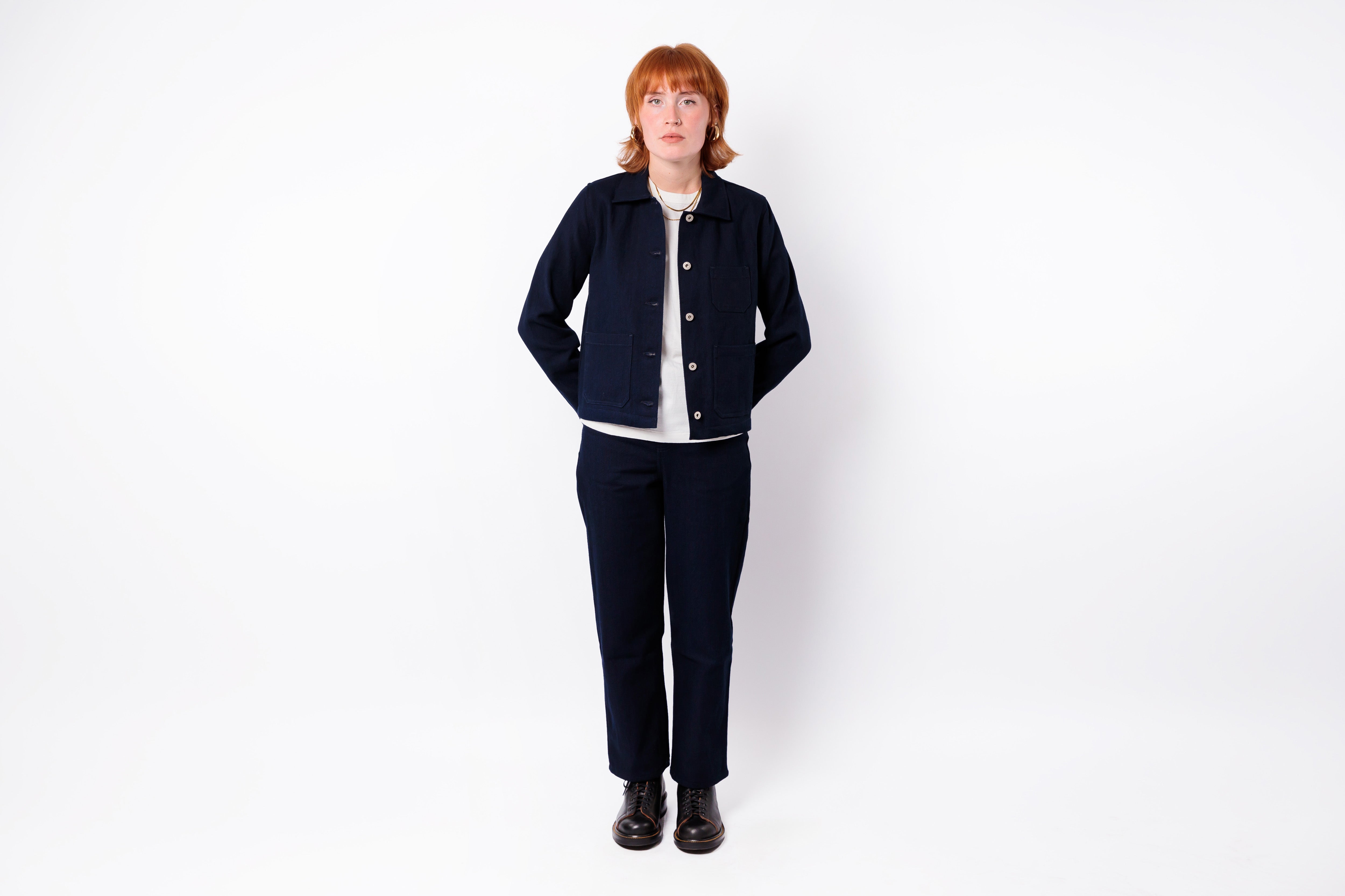 A model with short red hair stands against a plain white studio background wearing a dark navy blue jacket and matching trousers over a white shirt.