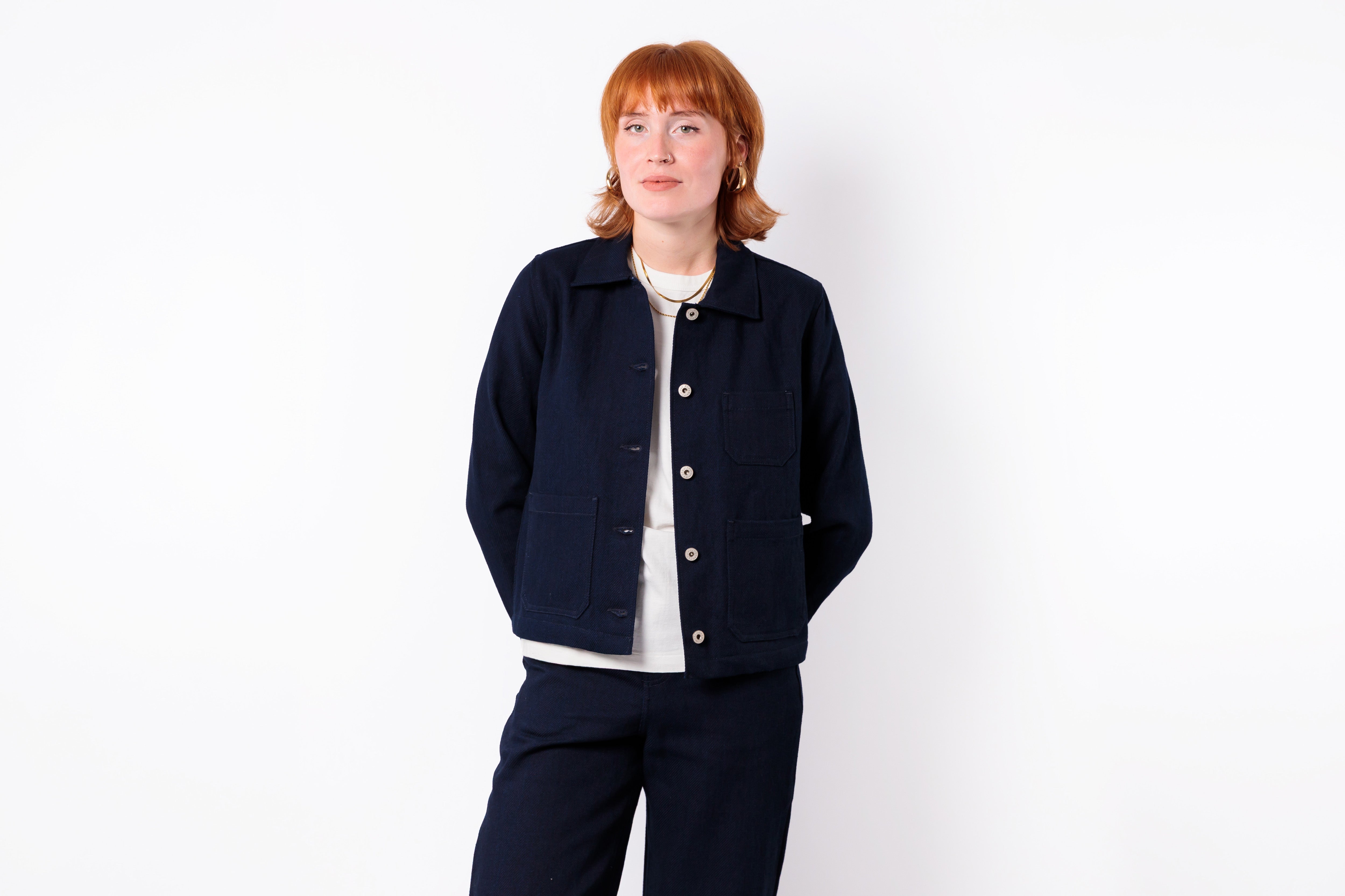 A woman with short red hair stands against a plain white background wearing a dark navy blue jacket over a white top and matching dark navy pants.