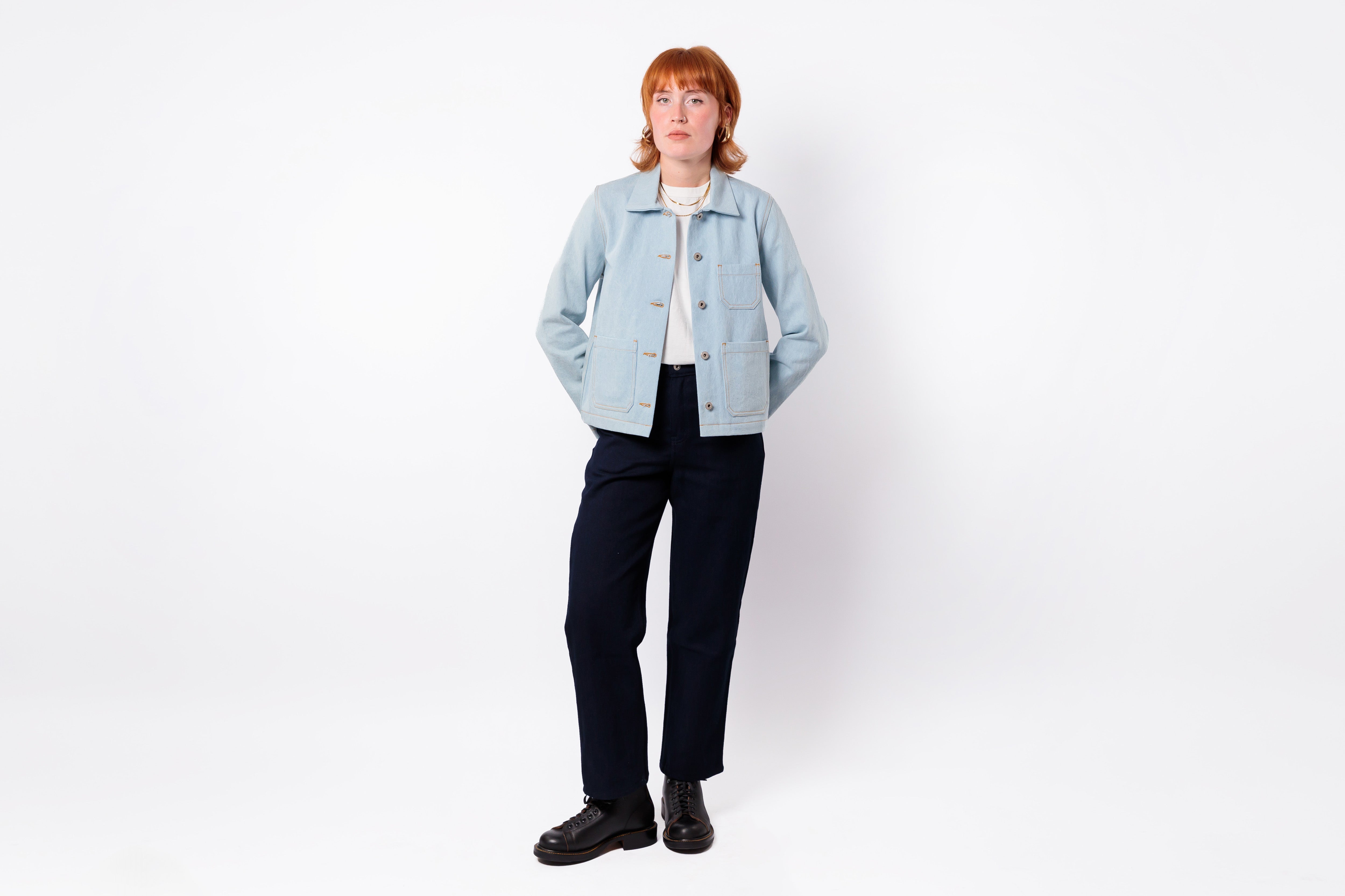 A model with short red hair stands against a white studio background wearing a light blue denim jacket over a cream turtleneck, dark navy trousers, and black shoes.