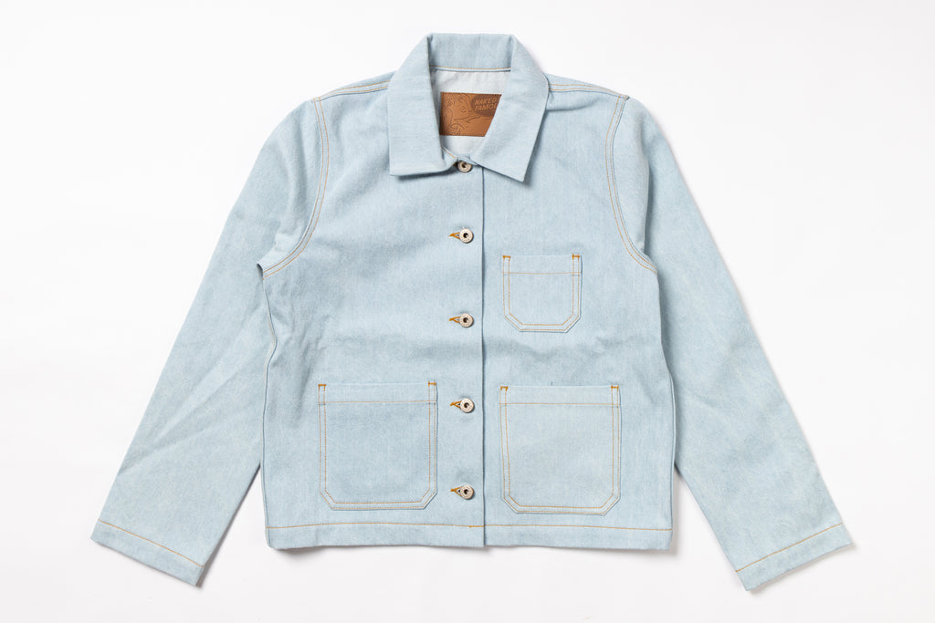 A light blue denim work jacket with a pointed collar, button closure, and four front pockets, laid flat against a white background.