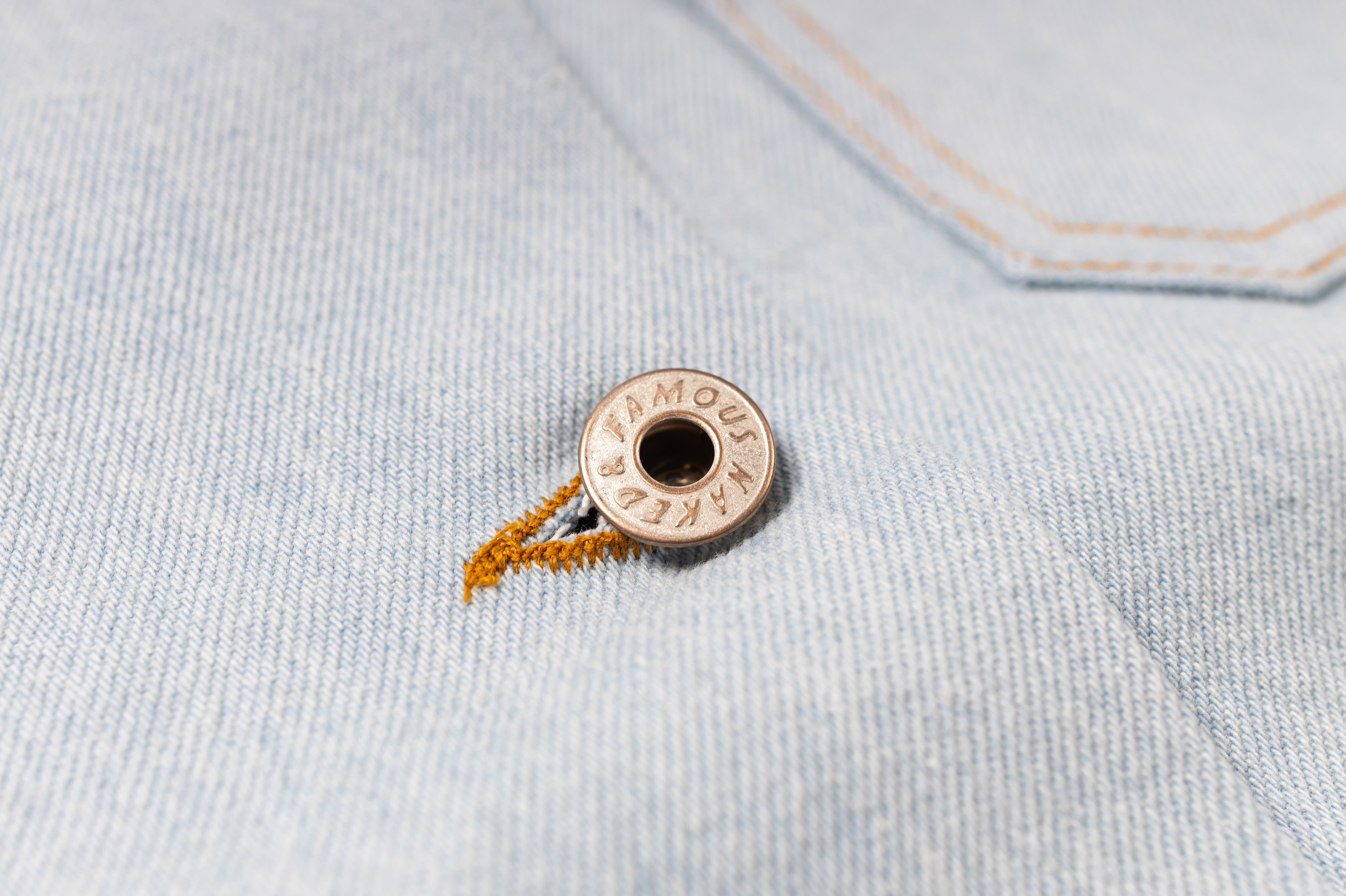 A close-up of a round metallic snap button with the brand name engraved on it, attached to a piece of orange thread, set against a light blue denim fabric background.