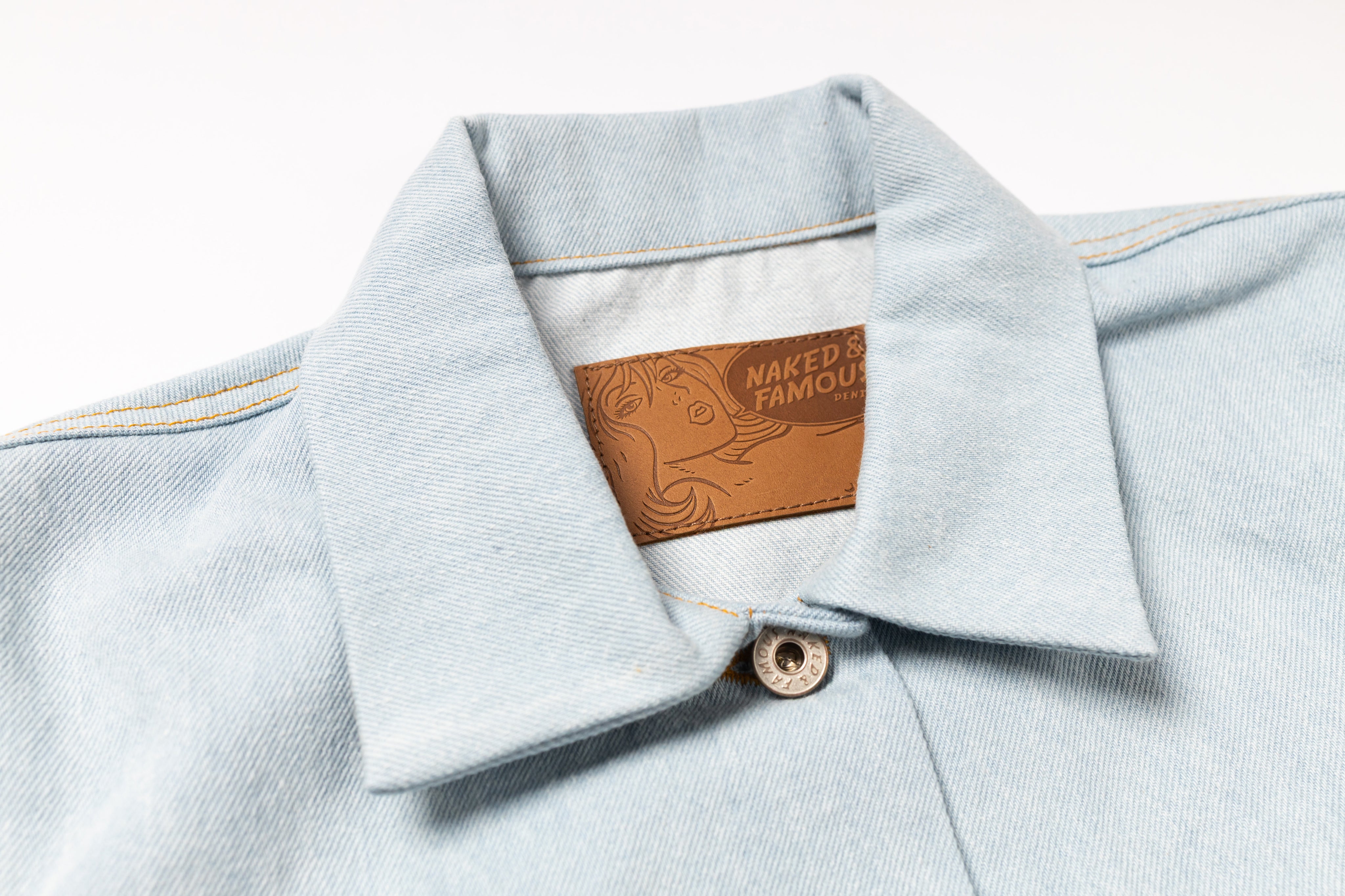 Close-up of a light blue denim shirt collar with a brown leather patch reading 'NAKED & FAMOUS' on a white background.