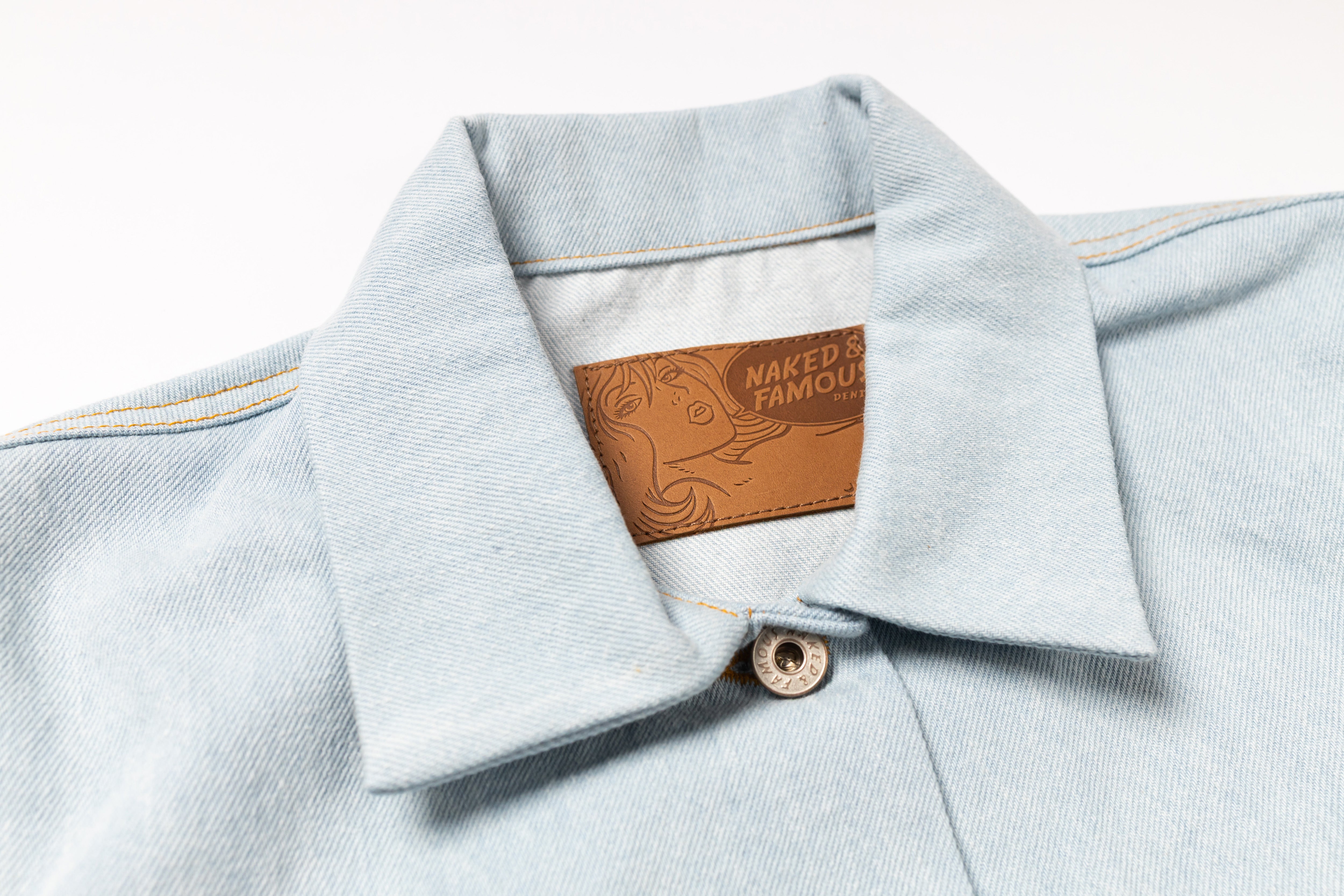 Close-up of a light blue denim shirt collar with a brown leather patch reading 'NAKED & FAMOUS' on a white background.