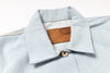 Close-up of a light blue denim shirt collar with a brown leather patch reading 'NAKED & FAMOUS' on a white background.
