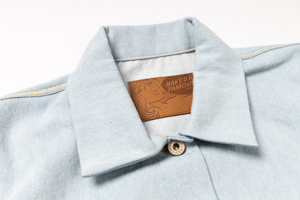 Close-up of a light blue denim shirt collar with a brown leather patch reading 'NAKED & FAMOUS' on a white background.