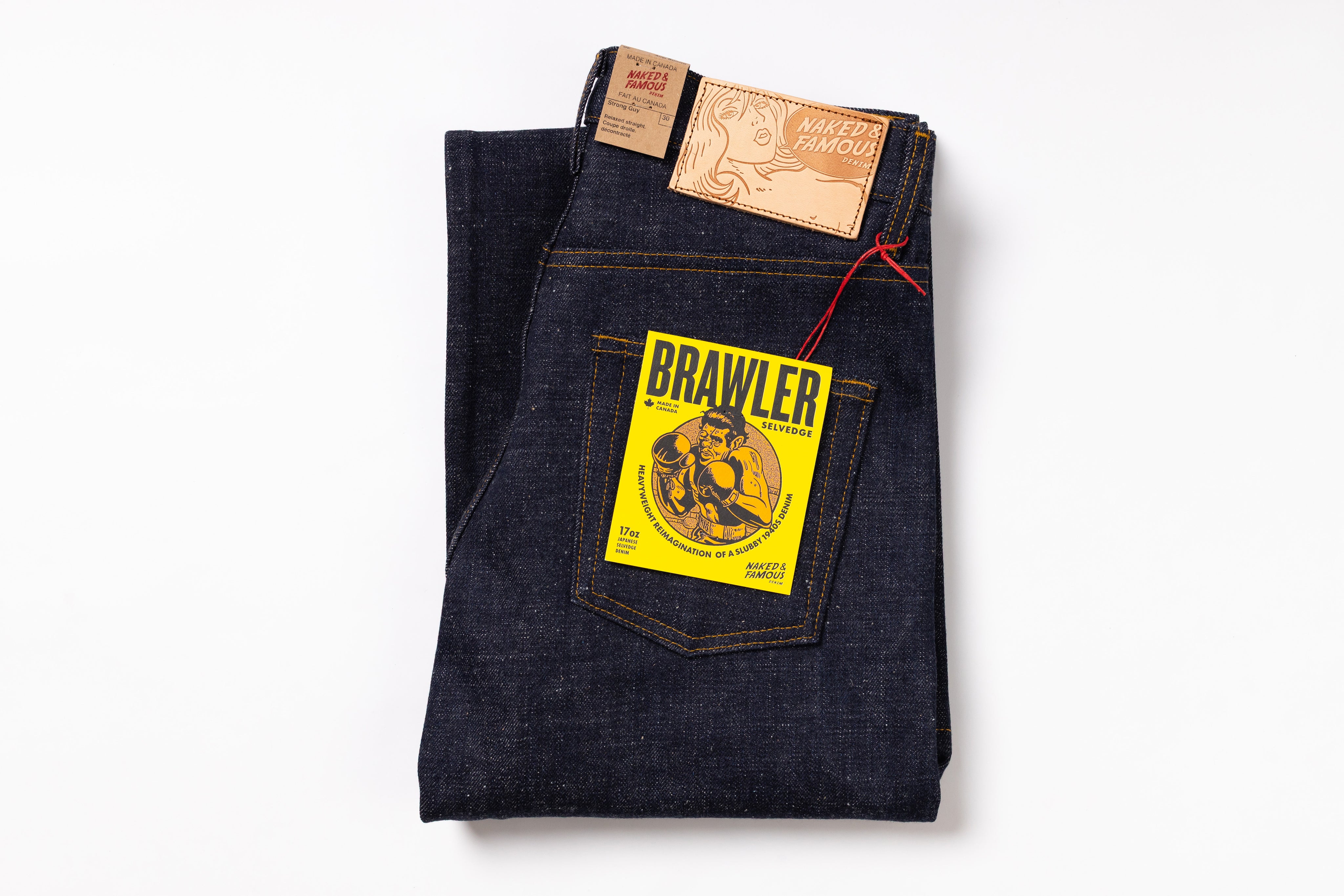 A pair of folded dark indigo denim jeans with a yellow Brawler brand patch on the back pocket, a leather brand patch at the waistband, and a red drawstring, photographed on a clean white background.