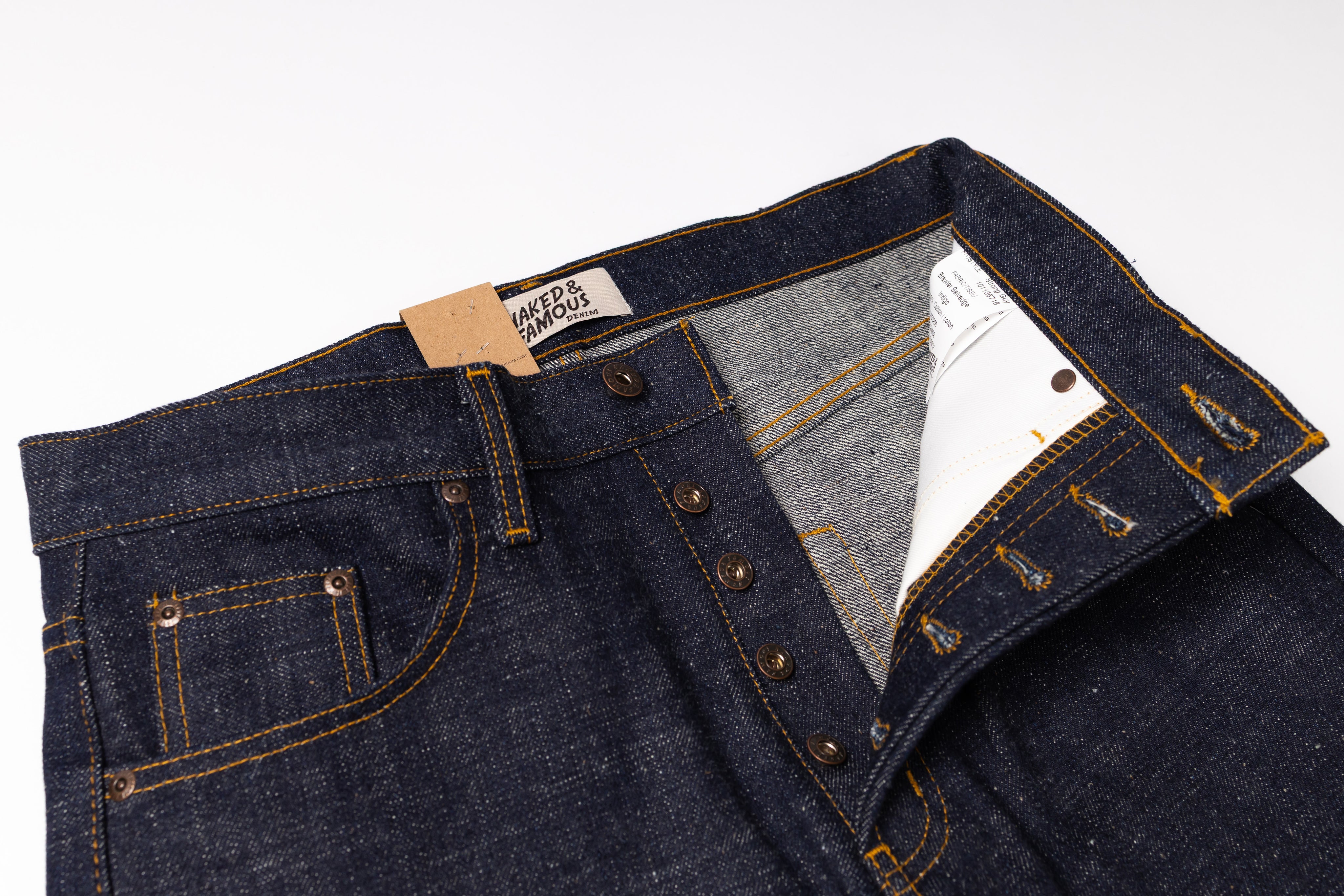 Close-up of a pair of dark indigo denim jeans showing the waistband, button fly, pocket, and interior lining with a paper tag attached.