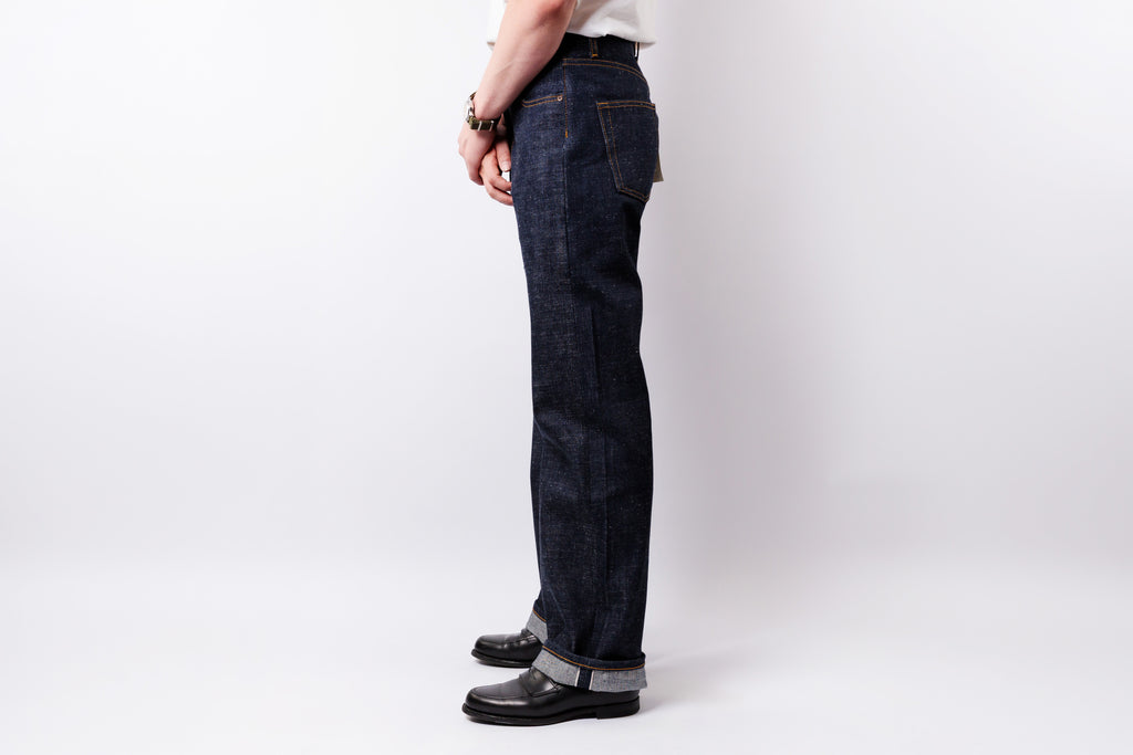 A person shown from the waist down wearing dark indigo denim jeans, black leather shoes, and a white shirt, standing against a light gray studio background.