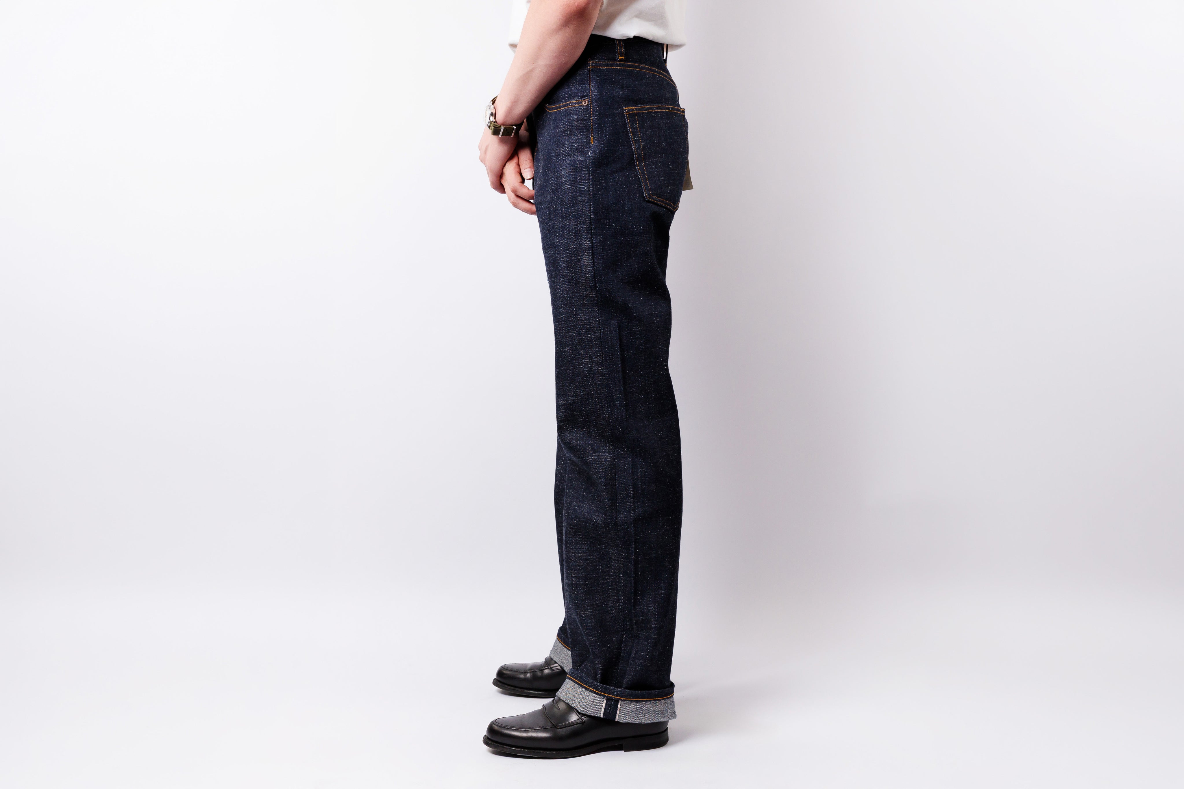 A person shown from the waist down wearing dark indigo denim jeans, black leather shoes, and a white shirt, standing against a light gray studio background.