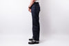 A person shown from the waist down wearing dark indigo denim jeans, black leather shoes, and a white shirt, standing against a light gray studio background.