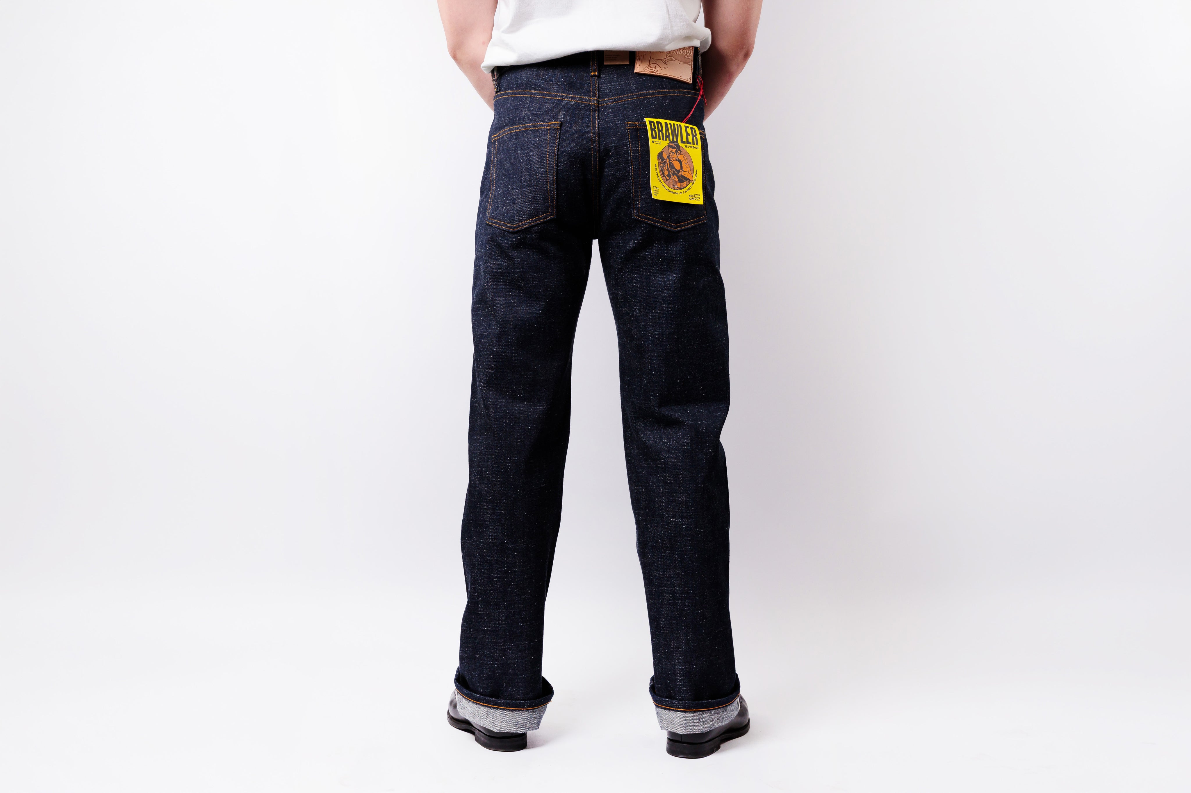 Rear view of a person wearing dark indigo denim jeans with a yellow and black patch on the back right pocket, paired with a white t-shirt and black shoes, against a plain white studio background.