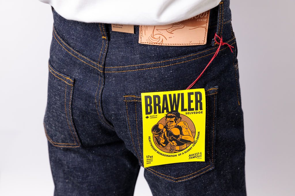 Close-up of the back waist of a person wearing dark indigo denim jeans with a yellow Brawler hangtag attached near the pocket, plus a white shirt hem visible at the top.