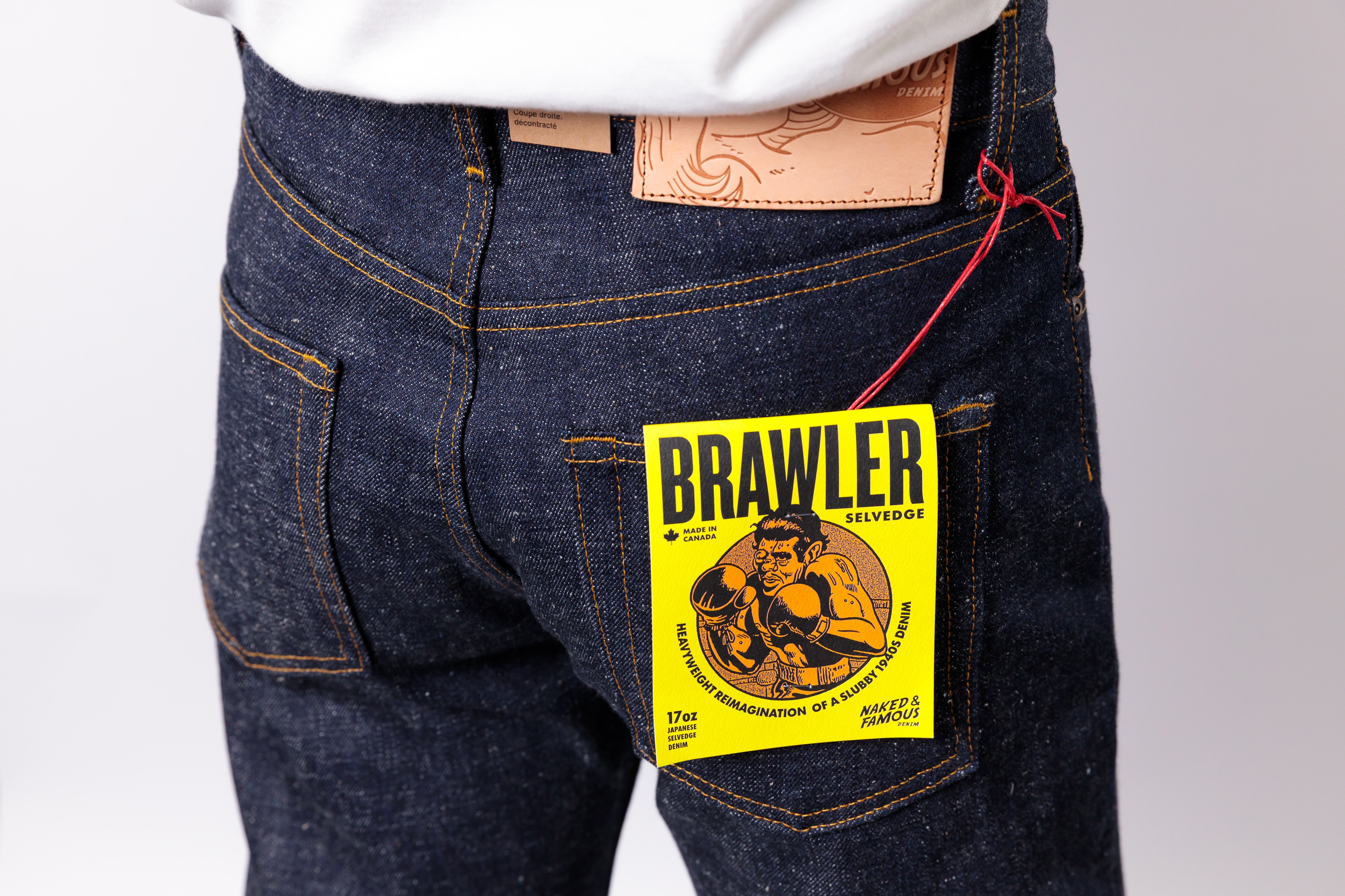 Close-up of the back waist of a person wearing dark indigo denim jeans with a yellow Brawler hangtag attached near the pocket, plus a white shirt hem visible at the top.