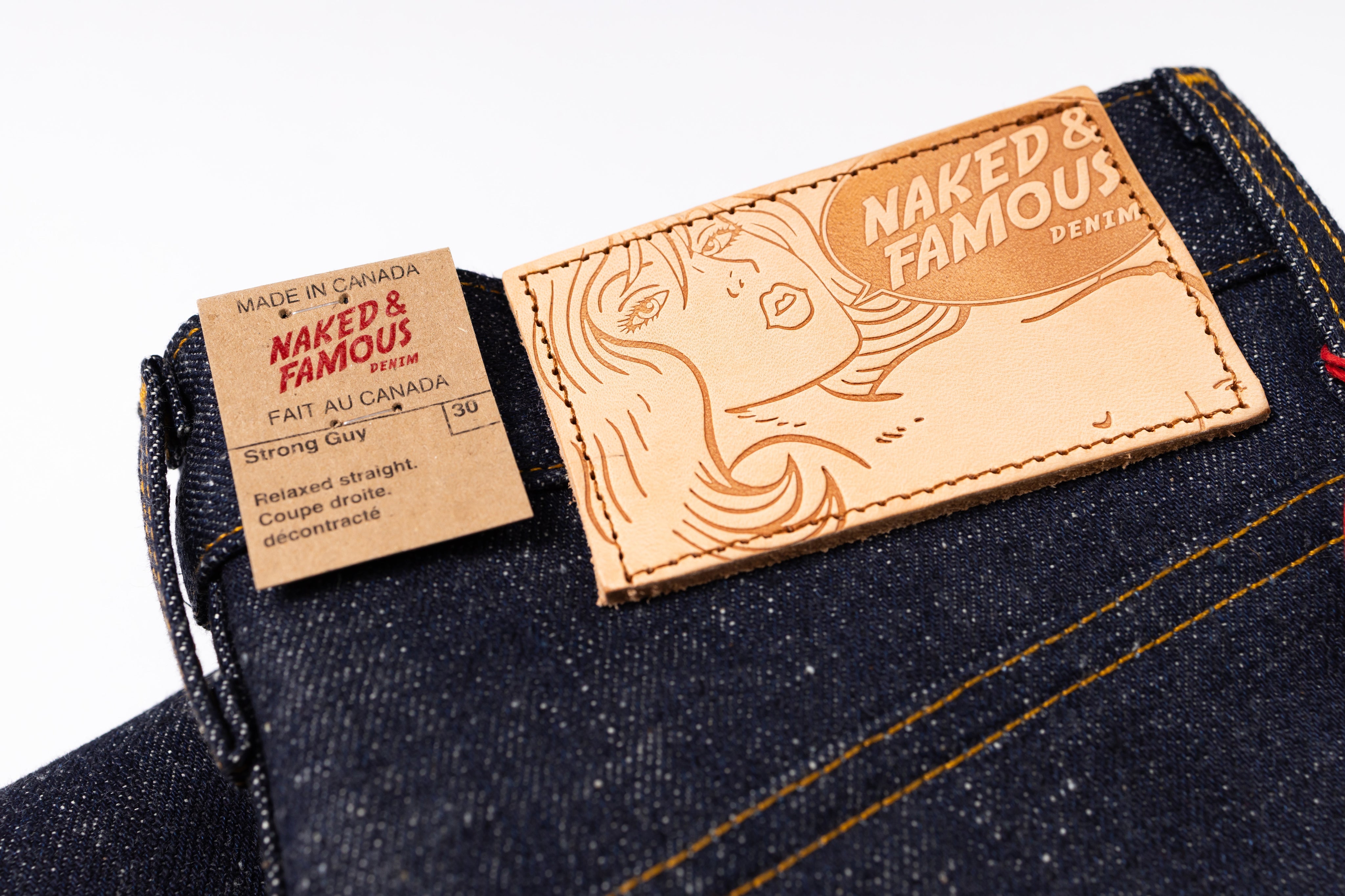 Close-up of the waistband of dark blue denim jeans showing two labels: a small brown paper tag reading 'NAKED & FAMOUS' and a larger tan leather patch with an engraved woman's face and the words 'NAKED & FAMOUS'.
