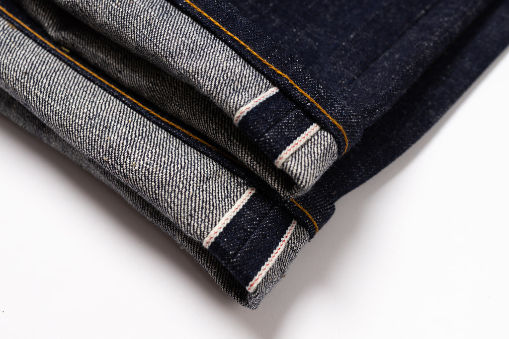 Close-up of folded dark indigo denim jeans showing the contrast tan/yellow contrast stitching and white stripe detail on the hem, set against a plain white background.