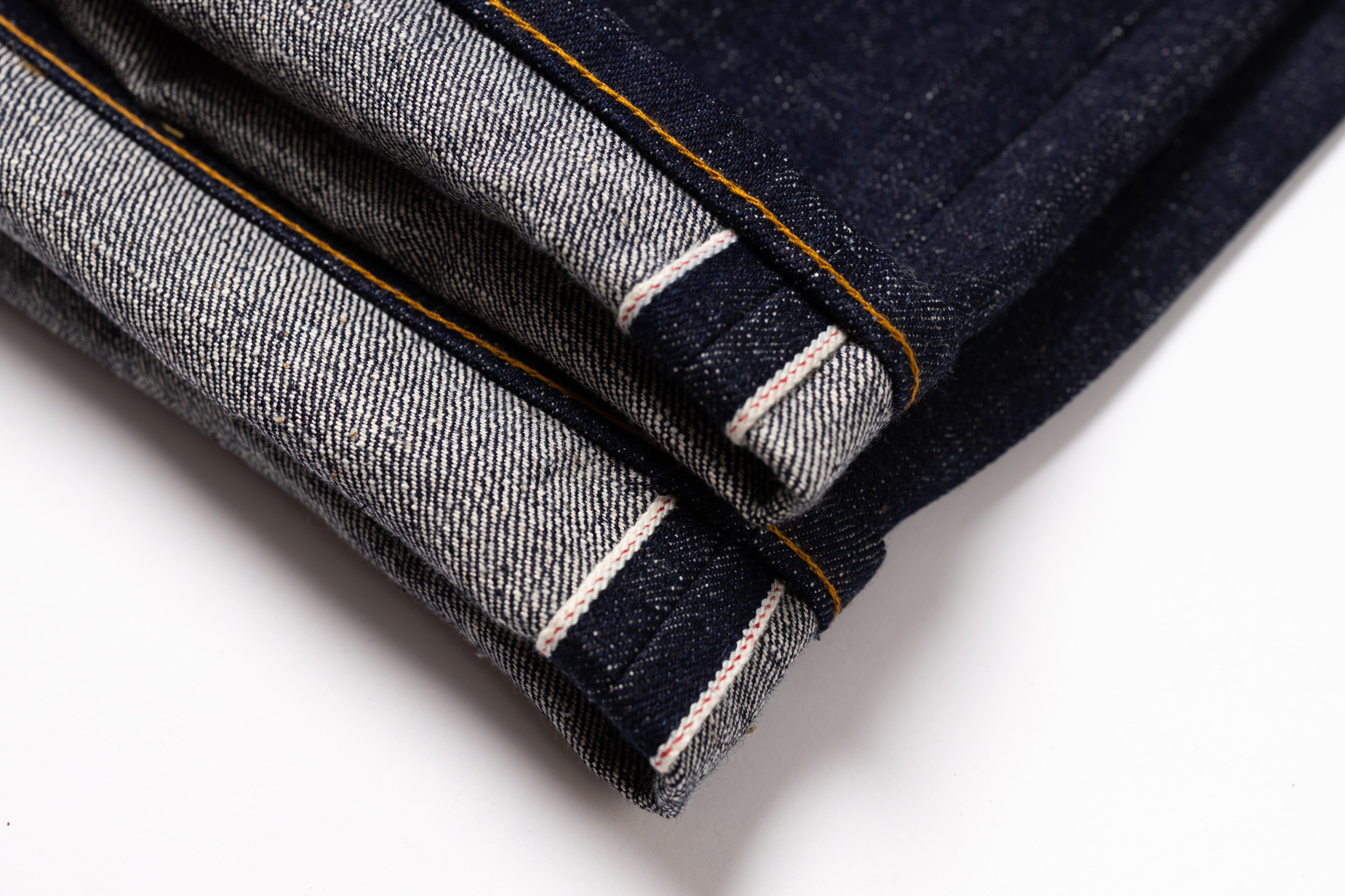 Close-up of folded dark indigo denim jeans showing the contrast tan/yellow contrast stitching and white stripe detail on the hem, set against a plain white background.