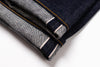 Close-up of folded dark indigo denim jeans showing the contrast tan/yellow contrast stitching and white stripe detail on the hem, set against a plain white background.